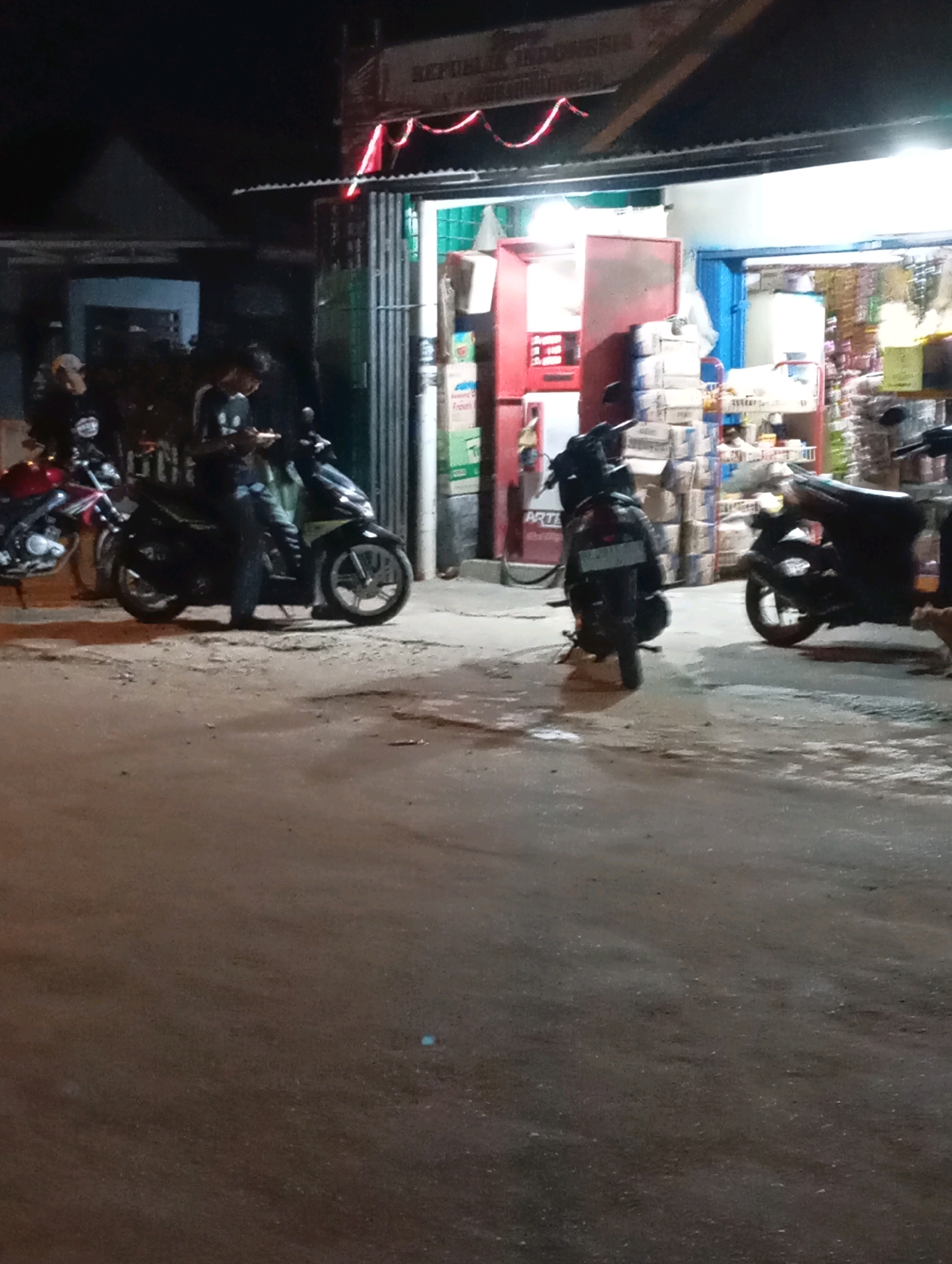 The image depicts a nighttime scene in front of a shop in Leman Benawi, Indonesia. Several motorcycles are parked on a paved area, with one person standing next to a motorcycle. The shop appears to be open, with goods displayed inside and stacked outside. Red and white decorative lights are visible above the shop entrance, and a sign with text is mounted on the building. The ground is a mixture of asphalt and dirt. The lighting is artificial, with the shop interior and some overhead lights illuminating the area. The overall atmosphere suggests a commercial street at night.
