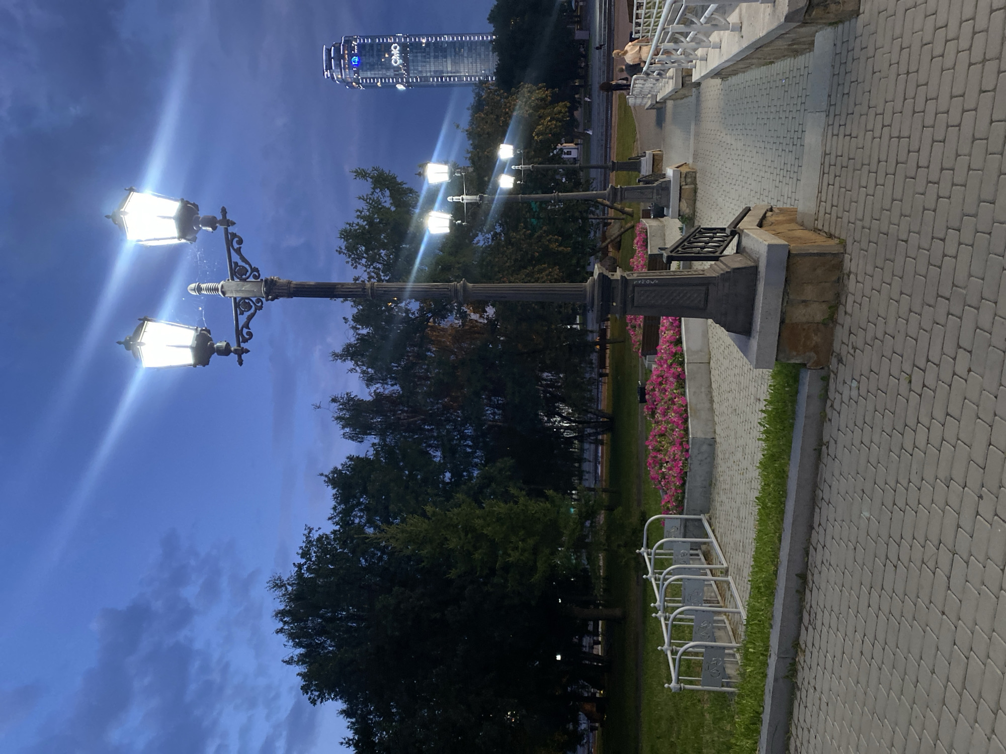 The image depicts a park area in Yekaterinburg, Russia, likely during the twilight hours, with a tall modern skyscraper illuminated in the background. A prominent streetlamp with multiple lights stands in the foreground, casting a bright glow. Adjacent to the lamp is a planter filled with vibrant pink flowers. A paved walkway with a decorative railing leads further into the park. In the middle ground, more streetlamps and trees are visible, creating a sense of depth. A few individuals are present, with two people walking along the path on the right side of the frame. The sky above is a deep blue with scattered clouds, suggesting the transition from day to night. The skyscraper in the background, identifiable by its illuminated "CMO" logo, is likely the Vysotsky Business Center.