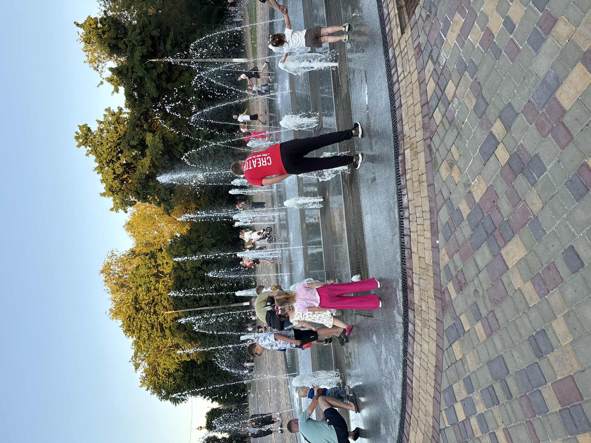 An outdoor fountain area in a city park is depicted, featuring multiple jets of water spraying upwards. Several people, including adults and children, are interacting with the fountain, some standing, some walking, and children are playing in the water. The foreground shows a paved area with a decorative stone pattern. In the background, lush green trees and a clear blue sky are visible. The scene appears to be set during a warm, sunny day, likely in the late afternoon or early evening given the lighting.