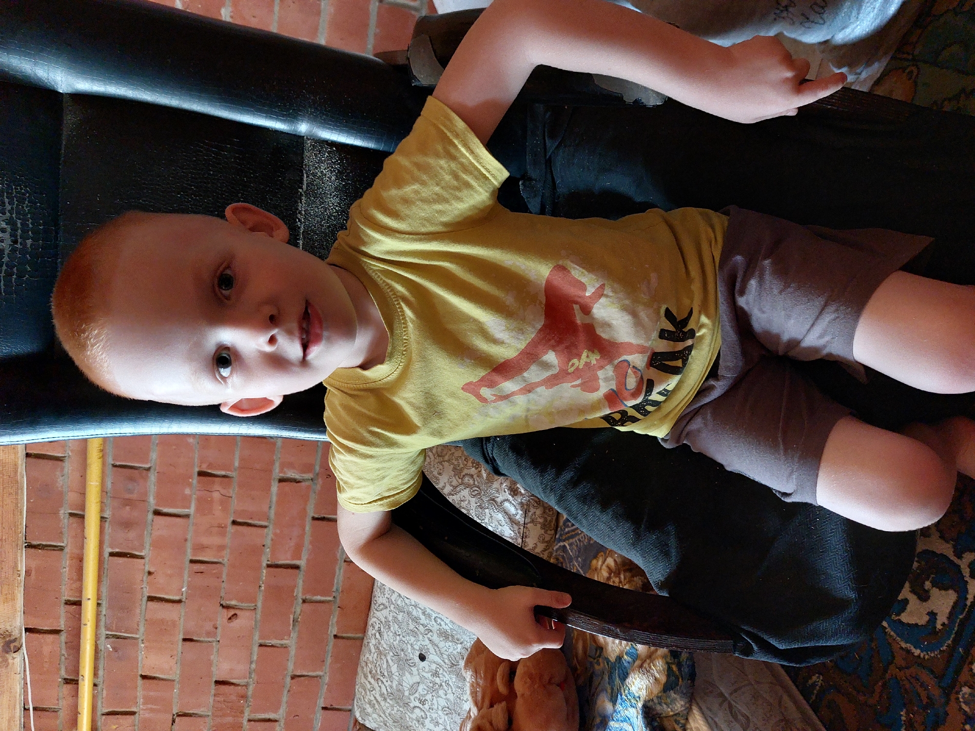 A young boy with light skin and reddish-blonde hair is lying on his back. He is wearing a yellow t-shirt with a graphic that appears to be a basketball player jumping, and gray shorts. The boy is positioned on a dark-colored surface, possibly a couch or chair, which is set against a brick wall. A yellow pipe runs vertically along the left side of the frame, against the brick. The floor beneath the boy is covered with a patterned rug. The overall impression is that of a casual indoor or semi-outdoor setting, possibly in a home. The boy's expression appears calm or slightly curious. There is no clear indication of the time of day or weather conditions.