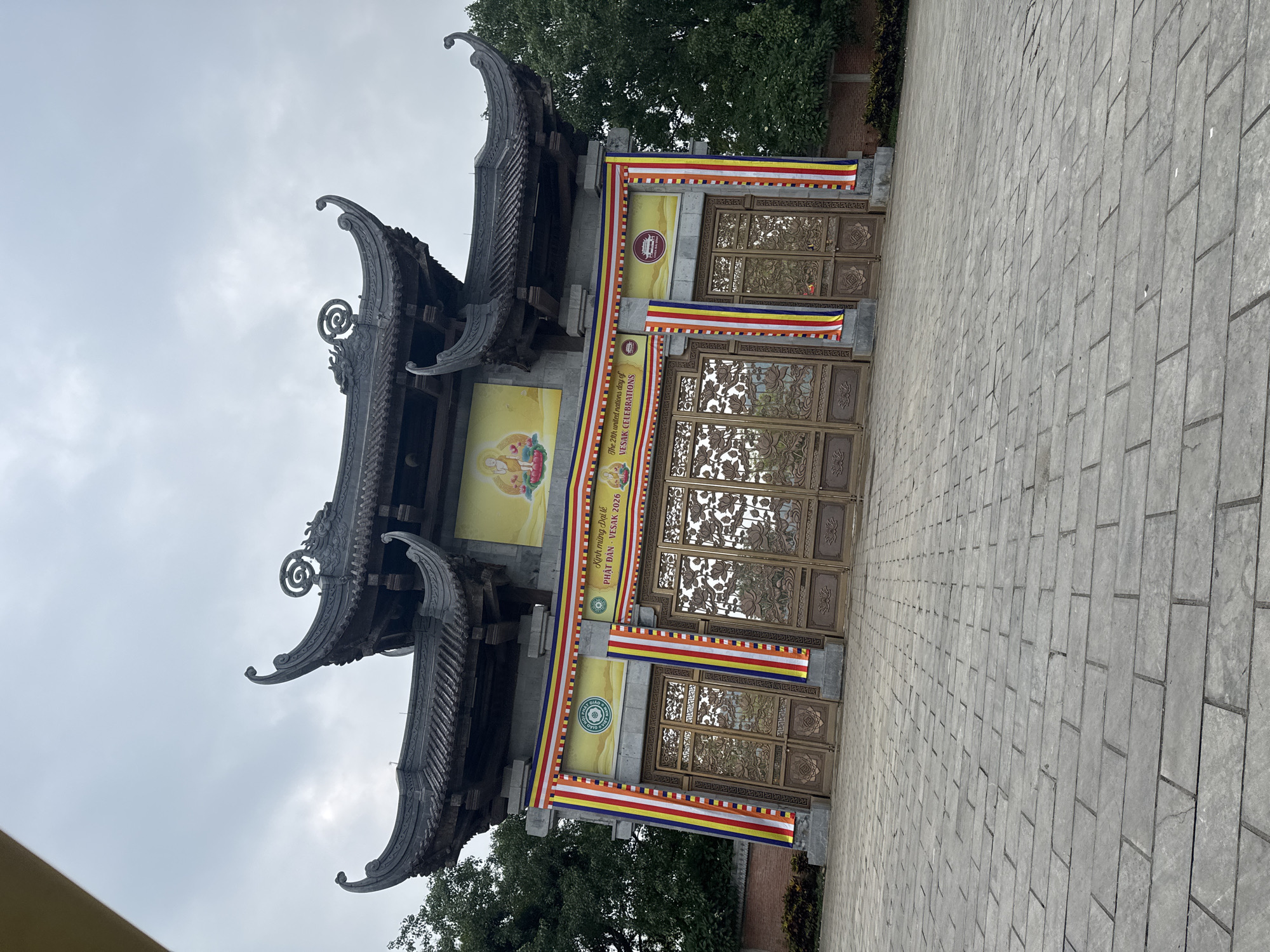 The image shows the entrance gate to a Buddhist temple in Tây Hoa Lư Ward, Vietnam. The gate is ornate, with a traditional East Asian architectural style, featuring curved roofs and intricate carvings. There are two large, golden gates with detailed lotus flower designs. Flanking the gates are banners with Buddhist imagery and text in Vietnamese and English, indicating a Vesak celebration. The ground in front of the gate is paved with large, grey stone tiles. Tall, green trees are visible on either side of the structure, and the sky is overcast. There are no people visible in the image. The overall impression is one of solemnity and grandeur associated with a religious site.
