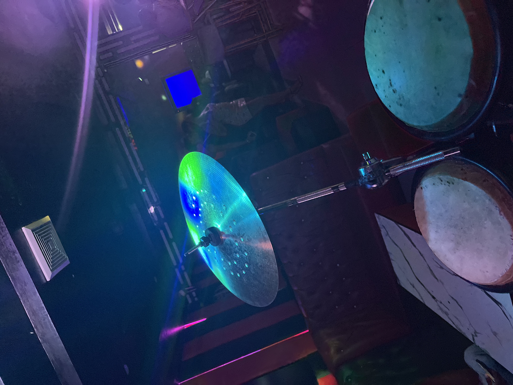 The image captures a close-up view of a cymbal and two drums in what appears to be a dimly lit entertainment venue. The cymbal, positioned prominently in the foreground and center, is illuminated with vibrant green and blue lights, reflecting an array of colors. It is mounted on a silver stand. To the right and left of the cymbal are the tops of two drums; the drum on the right is illuminated with a teal light, and the one on the left is a lighter, muted tone.
In the background, the scene is blurred and darker, suggesting the presence of people and a lively atmosphere. Vertical red and black striped patterns are visible on the left side of the background, possibly indicating a wall or stage decor. Above, a dark ceiling features a rectangular vent and metallic beams. A blue screen or monitor is visible in the mid-background, and the overall lighting suggests a club or concert setting with colorful stage lights casting shadows and highlights throughout the space. The location is described as Unknown, Unknown.