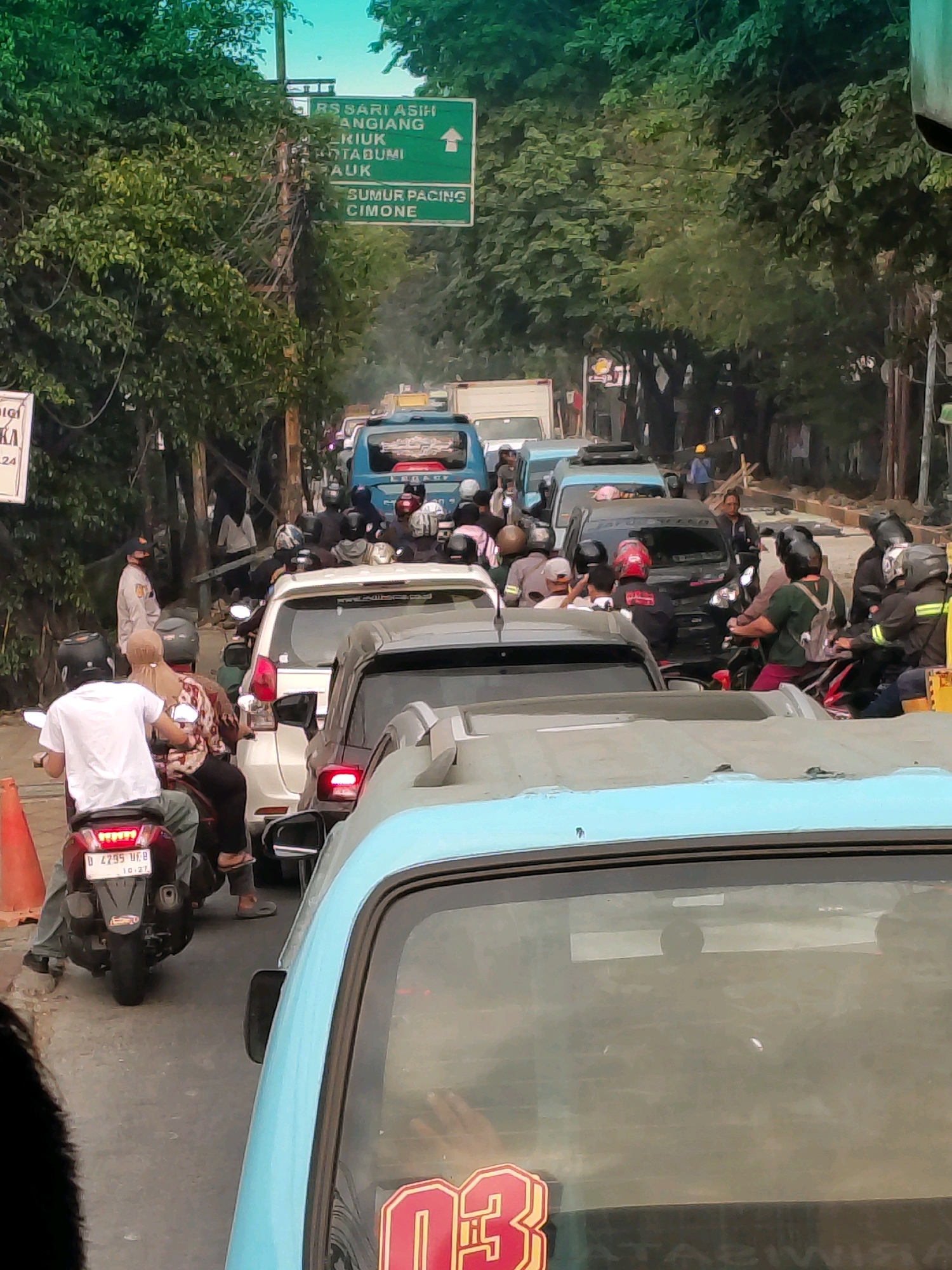 This is a vertical, eye-level, slightly tilted, medium shot of a congested road in Tangerang, Indonesia, likely during daytime, with a clear sky visible at the top. The image captures a traffic jam with numerous vehicles, including cars, motorcycles, and a blue bus. Several people are on motorcycles, wearing helmets. One person in a white shirt is on a motorcycle in the foreground, and another person in a patterned top is on the same motorcycle behind them. A man in a light-colored uniform stands near a utility pole. A green street sign indicates directions to "RS SARI ASIH," "ANGIANG," "STADUM," "PAUK," "SUMUR PACING," and "CIMONE." Trees line the sides of the road, and various poles and wires are visible. The front of a light blue vehicle dominates the bottom of the frame, obscuring the road ahead.
