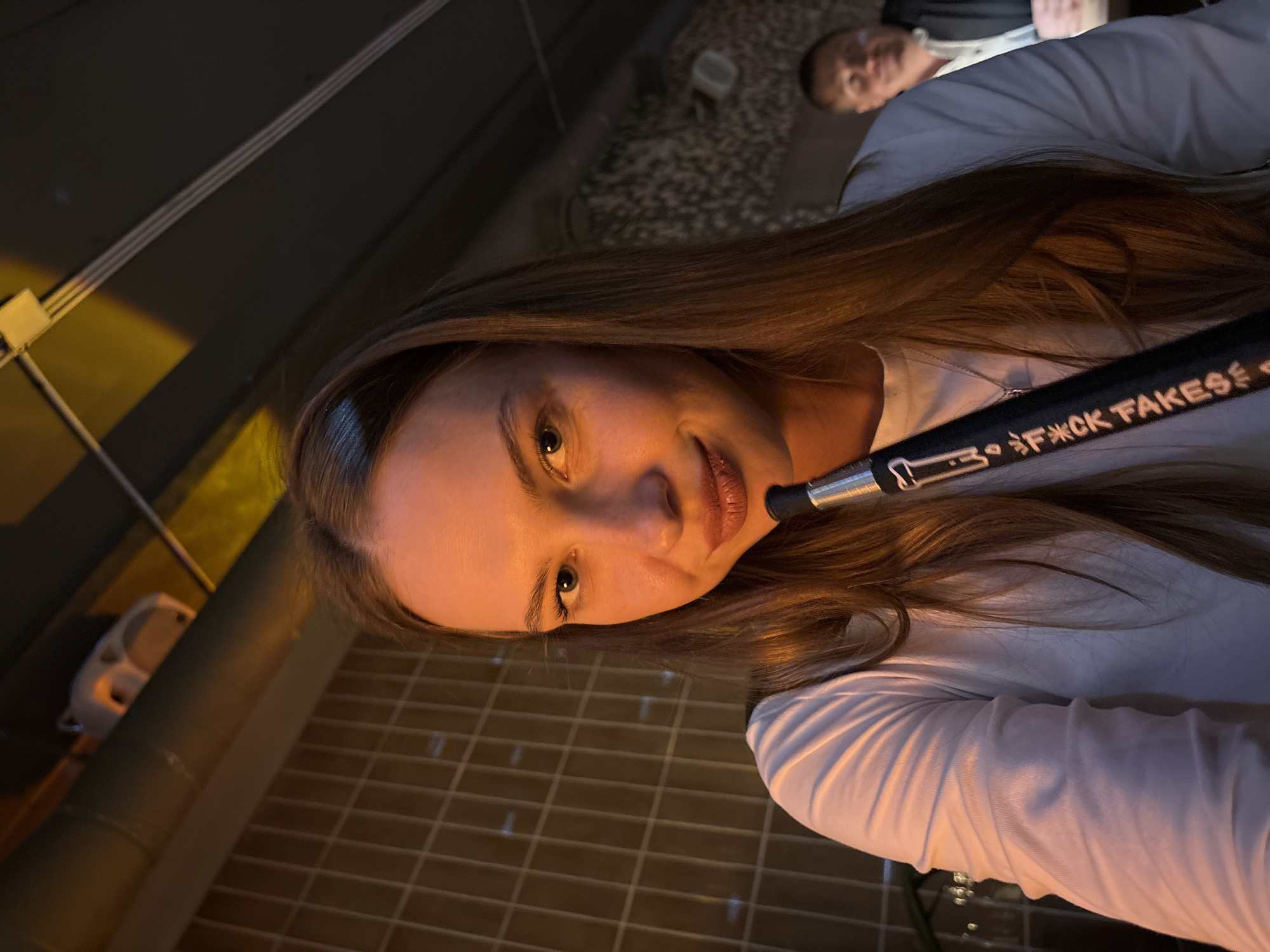A young woman with long, light brown hair is shown in a close-up shot, holding a black hookah hose with white lettering that reads "fuck fakes". She is wearing a white long-sleeved shirt and a silver pendant necklace. Her face is illuminated by warm lighting, highlighting her blue eyes and neutral expression. The background suggests an indoor setting with a tiled wall to the left and a dimly lit area with a blurred male figure in the background to the right. There are also exposed pipes and ceiling fixtures visible. The overall ambiance is casual and social, potentially a hookah lounge or bar. There are no distinct landmarks to identify the city as Perm, Russia.