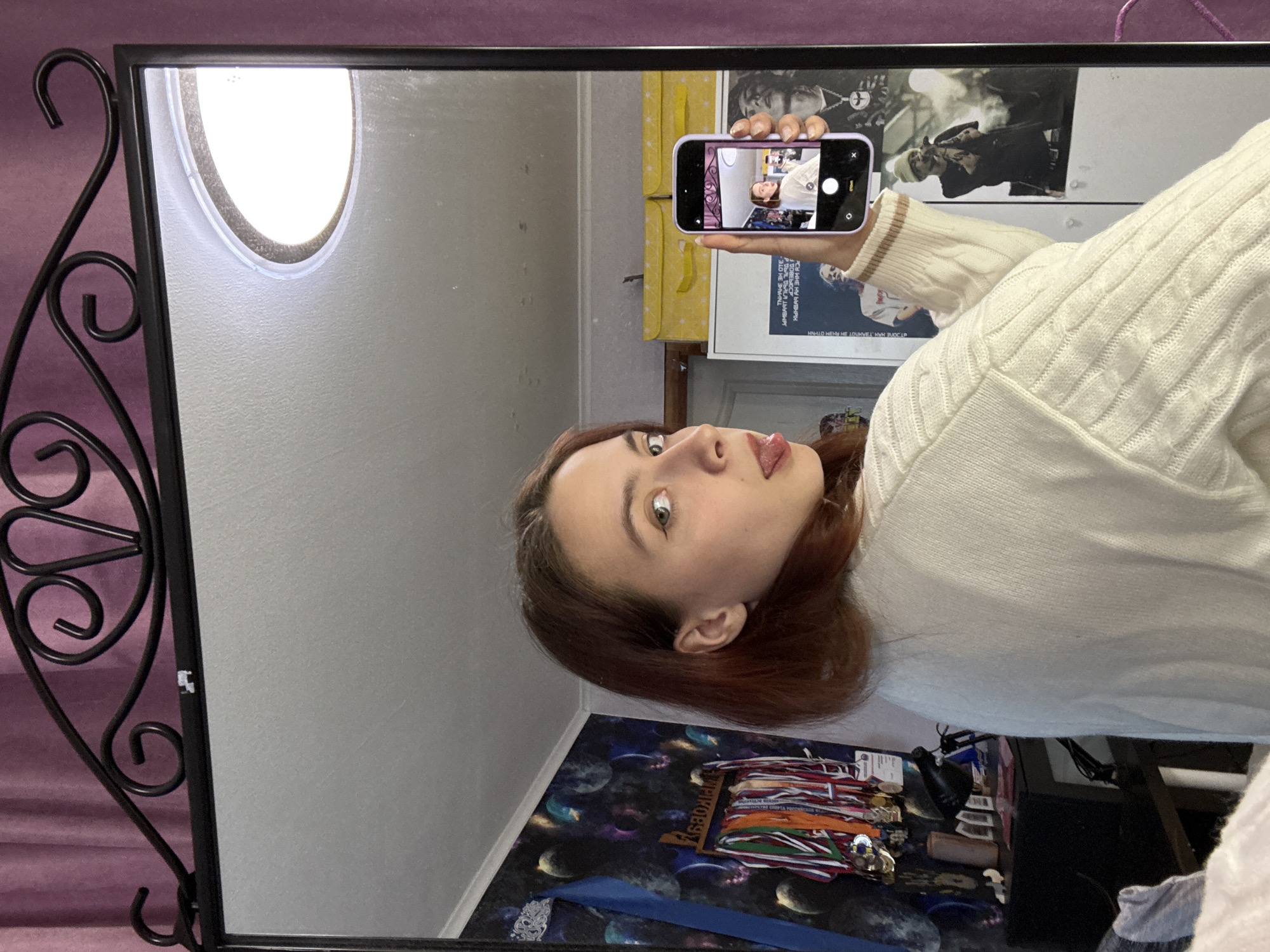 Here's a detailed description of the image:
The image shows a person taking a selfie in a mirror. The person, likely female, has auburn hair and is wearing a white sweater with a cable knit pattern. She's sticking her tongue out in a playful expression.
The person is holding a smartphone, which is also reflecting in the mirror. The phone's screen shows a miniature version of the selfie being taken. The phone has a light purple/lavender case.
The mirror has a black ornate metal frame with decorative swirls and curves. The background visible in the mirror includes a poster featuring black and white images of people, a yellow patterned curtain, and a light-colored wall.
Below the person, there's a cluttered surface with a space-themed pattern. Various objects are visible on the surface, including what appear to be medals or ribbons and other miscellaneous items.
The image also shows a small sticker on the black metal frame of the mirror.
