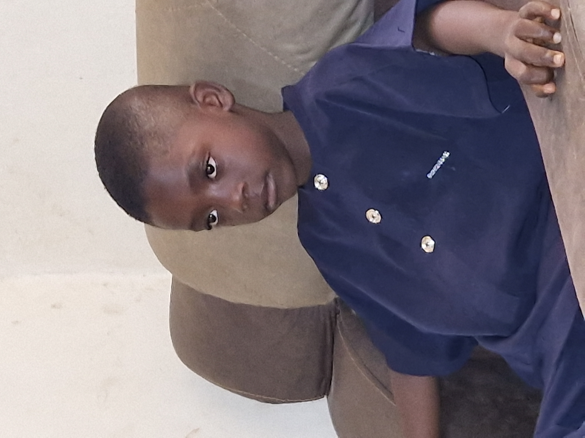 A young boy with dark skin and short hair is seated on a brown chair. He is wearing a dark blue shirt with decorative buttons. The background is a plain wall.