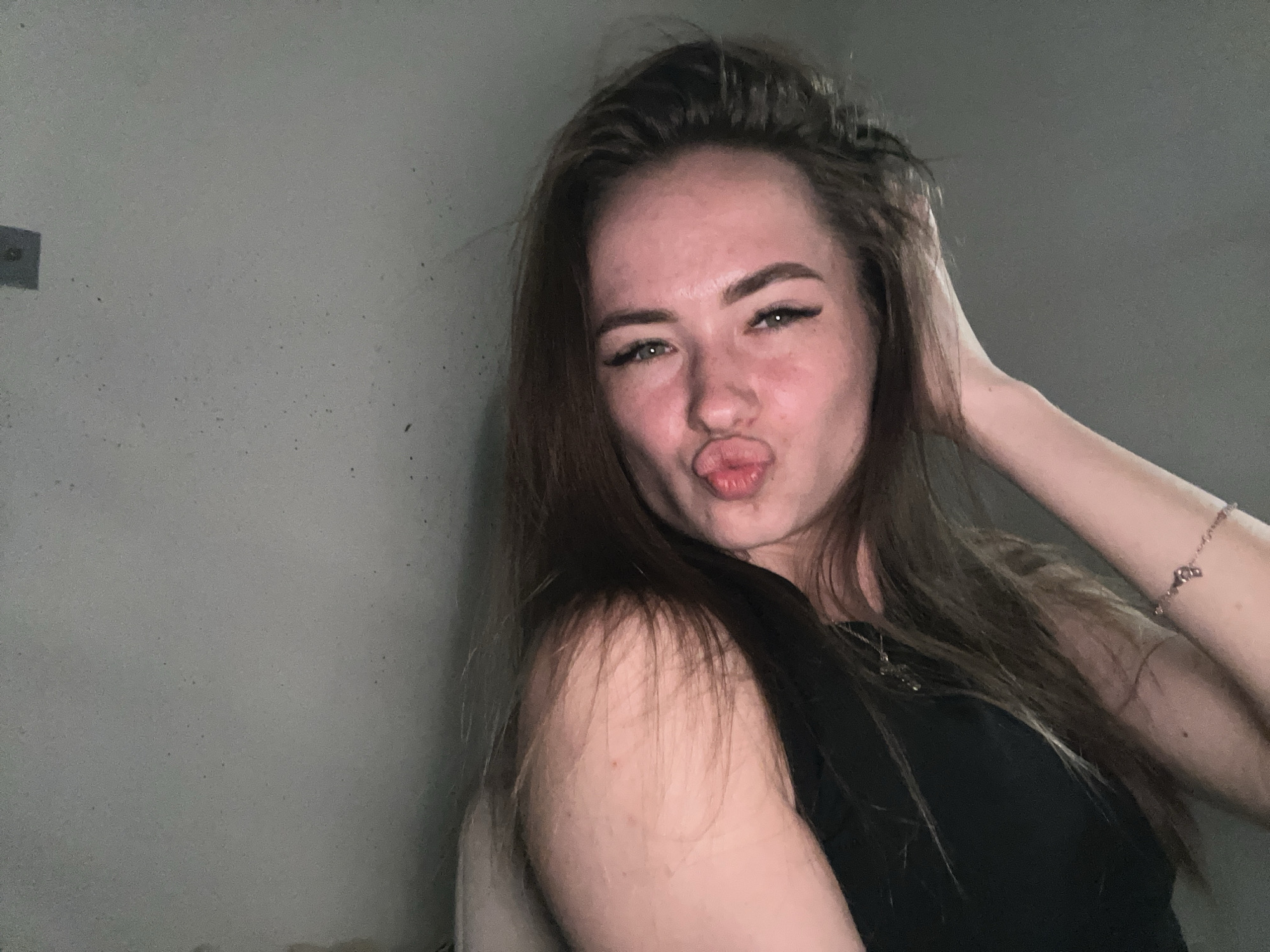 A close-up, chest-up portrait depicts a young adult female in the city of Unknown, Unknown. The subject, positioned slightly right of center, has fair skin exhibiting some diffuse redness on the cheeks and forehead. Her long, dark brown hair is styled loosely, extending over her shoulders. She is looking directly at the camera with a "kissing face" expression, lips pursed into a pout. Her eyes, appearing green or hazel, feature prominent black winged eyeliner. Her left hand (viewer's perspective) is raised, with fingers partially obscured by her hair, touching the back of her head. A delicate, light-colored bracelet with small charms is visible on her left wrist. She is wearing a dark, possibly black, sleeveless top or tank top, with a faint metallic chain visible on her chest. The background consists of a plain, light grey wall with minor texture and small, scattered dark speckles. A dark, rectangular object is partially visible on the upper left section of the wall. The lighting appears artificial, highlighting the subject's face and creating subtle shadows.