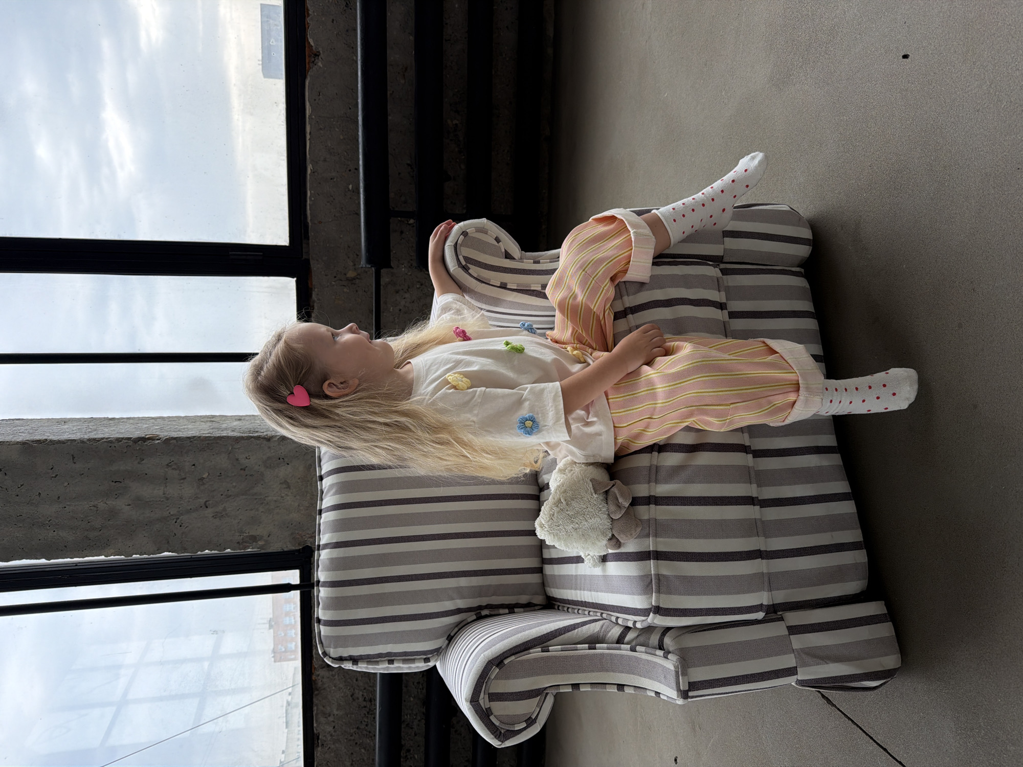 A blonde-haired girl lies on a striped grey and white chair. She wears pink and yellow striped pajamas and white socks with red polka dots. She has a pink heart clip in her hair. Next to her is a small plush toy resembling a sheep. The chair is positioned near a large window with black frames, suggesting an industrial or loft-style interior. Outside the window, cloudy sky is visible, likely indicating a time of day when the sun is not directly shining. The floor is a neutral grey, possibly concrete. The background includes elements of architecture, suggesting an urban setting, but there is not enough information to identify the location in the city of Saratov, Russia.
