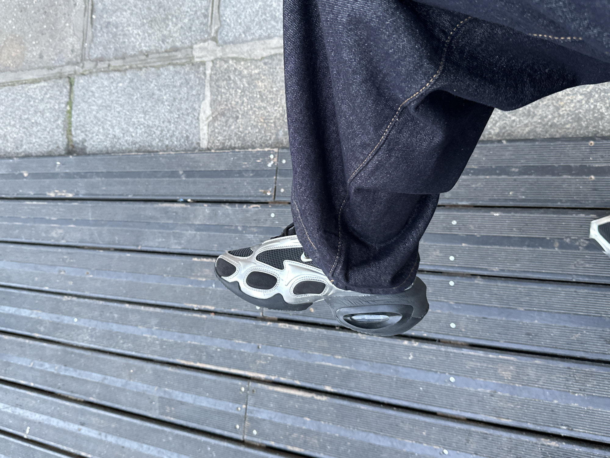 A medium shot captures a person's lower body, specifically the left foot wearing a silver and black athletic shoe. The person is clad in dark denim pants. The subject is on a wooden boardwalk or walkway, with the boards running horizontally. To the left, a section of gray stone or concrete tiles is visible. The scene is nondescript and lacks distinct features or landmarks that would definitively place it in Paris, France.