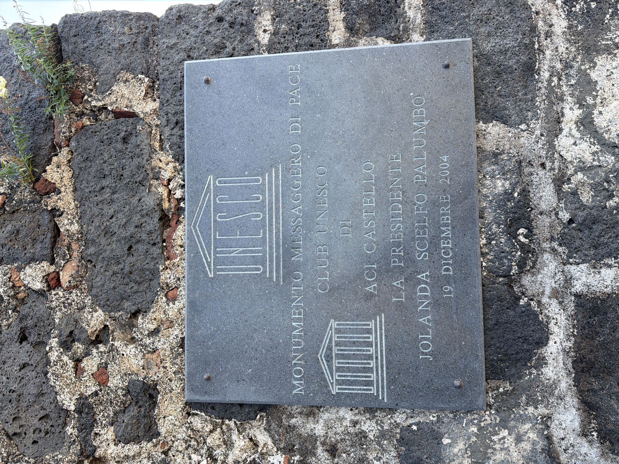 The image shows a gray stone plaque mounted on a dark stone wall. The plaque is inscribed with the word "UNESCO" above the words "MONUMENTO MESSAGGERO DI PACE" and "CLUB UNESCO DI ACI CASTELLO." Underneath these lines are the words "LA PRESIDENTE JOLANDA SCELFO PALUMBO" followed by "19 DICEMBRE 2004". The image indicates that the plaque is a monument in Aci Castello, Italy.