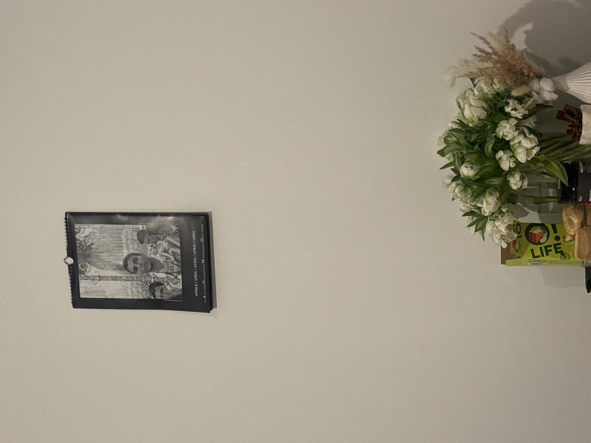 The image presents an indoor scene with a light-colored, possibly off-white or beige, wall as the dominant background. The composition is split, featuring a calendar on the left and a floral arrangement on the right.
On the left side of the frame, a black-bound calendar is visible. It features a black and white photograph depicting what appears to be a group of people. A man with dark hair is clearly visible in the center, looking towards the camera, with other figures partially obscured behind him. The photograph's aesthetic suggests a vintage or retro style. Below the image, calendar text is visible, including "APRILE / APRIL / AVRIL / ABRIL" (indicating the month of April in several languages) and a sequence of dates, with numbers such as "1, 2, 3, 4, 11, 18, 25, 29" discernable. A white hook or tab is attached to the top spiral binding, suggesting how it is hung.
On the right, a vibrant floral arrangement sits on a surface, likely a shelf, casting a soft shadow on the wall above. The bouquet consists of numerous white flowers with green foliage, possibly artificial tulips, arranged in a clear glass vase. Complementing the white flowers are dried decorative elements, including fluffy pampas grass and white cotton-like clusters, displayed in or alongside a ribbed white vase. Next to these, a brown, textured decorative object is partially visible. Below the flowers and vases, a green box or package is seen, bearing the prominent white text "O! LIFE" accompanied by a red exclamation mark. Below this, the word "CREATIVE" is faintly visible.
The scene depicts a static interior arrangement with no human activity taking place. The lighting suggests an indoor setting, possibly with artificial illumination. No specific time of day or weather is indicated. The overall impression is one of simple home decor.