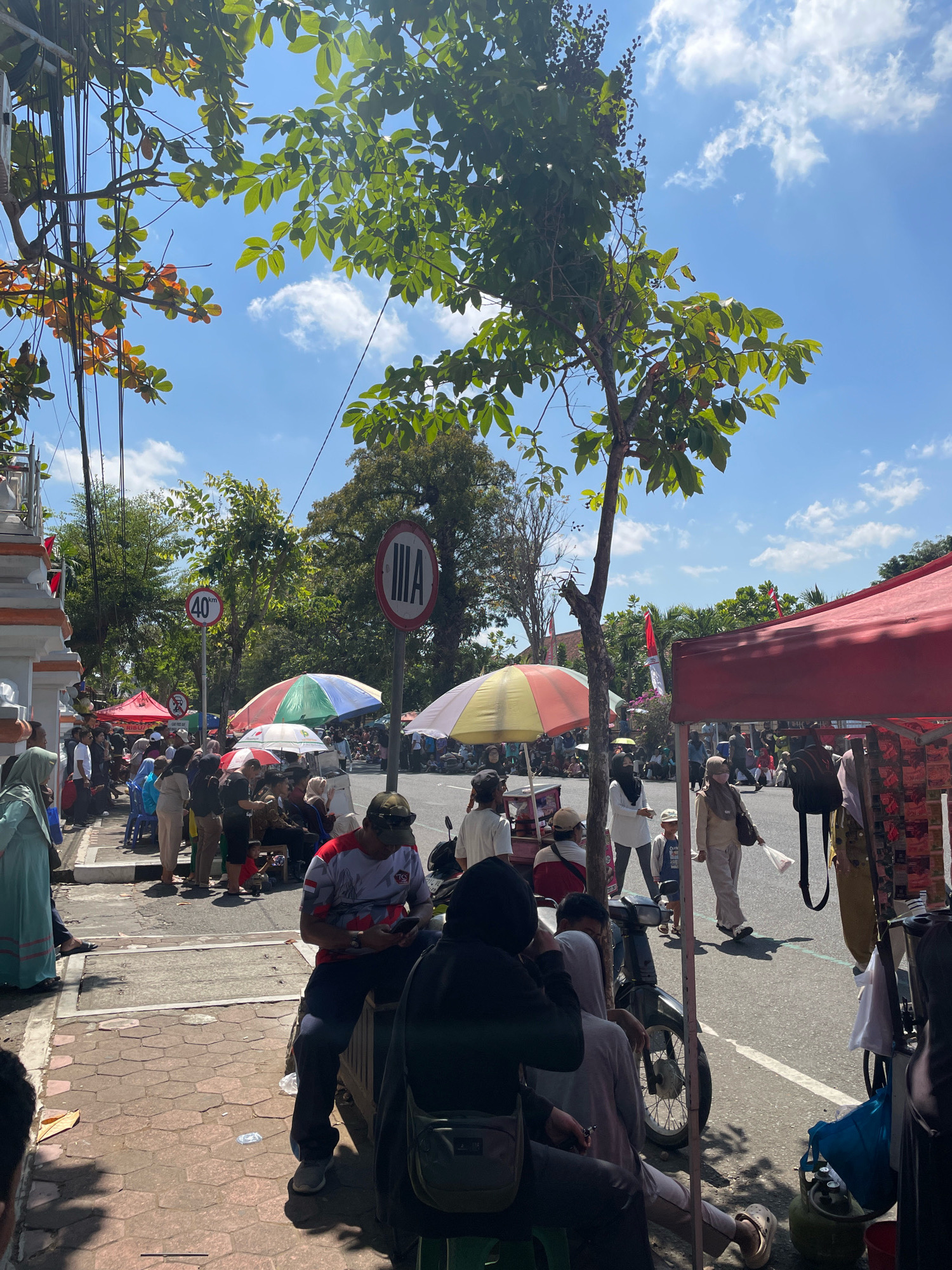 The image depicts a bustling street scene in Purworejo, Indonesia, under a bright blue sky with scattered clouds. A large tree with green leaves shades a portion of the sidewalk. On the sidewalk, people are gathered, some sitting on a low wall, others standing and walking. They appear to be observing something or waiting. Various stalls and tents are set up along the street, suggesting a market or a local event. One stall features a red canopy displaying merchandise.
In the background, a road is visible with vehicles and more people. Traffic signs are present, including a speed limit sign indicating 40 km/h and another sign with a circular red border and a vertical bar inside, also in a circular red border. Several colorful umbrellas are deployed over some of the stalls, providing shade to vendors and customers. The overall atmosphere is lively and suggests a public gathering.