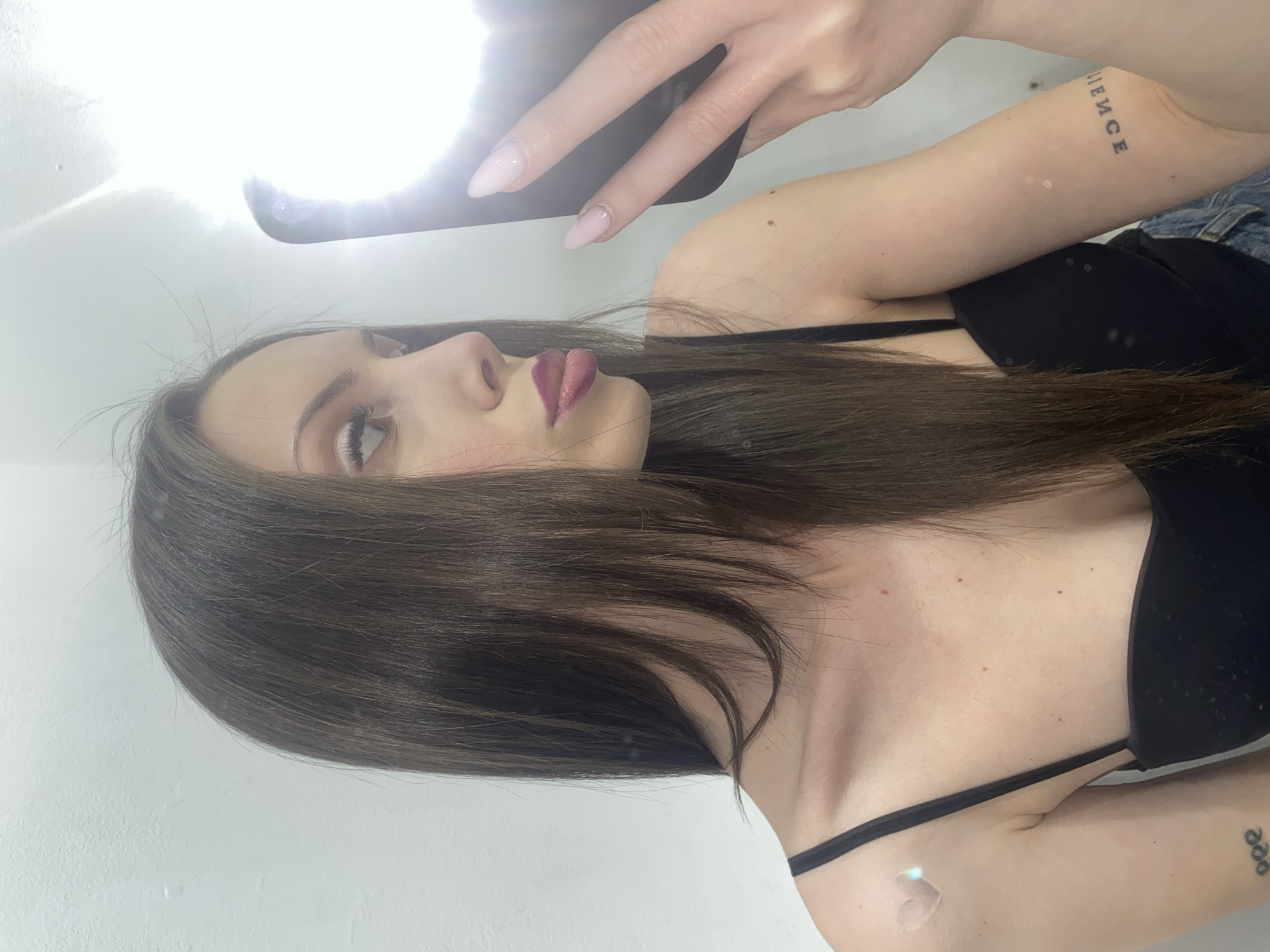 A young woman with long, dark brown hair and light makeup is taking a selfie in a well-lit room with white walls. She is wearing a black camisole and appears to be looking at her phone, which is held in her right hand, with the flash directed towards her face. She has a light pink lipstick on and her tongue is slightly sticking out. There are a few small blemishes visible on her skin, and she has a small heart-shaped tattoo on her left shoulder and an arm tattoo that reads "LIENCE" in reverse on her right arm. The background is a plain white wall, reflecting the light from the phone. The overall impression is a casual, personal moment captured by the woman herself.