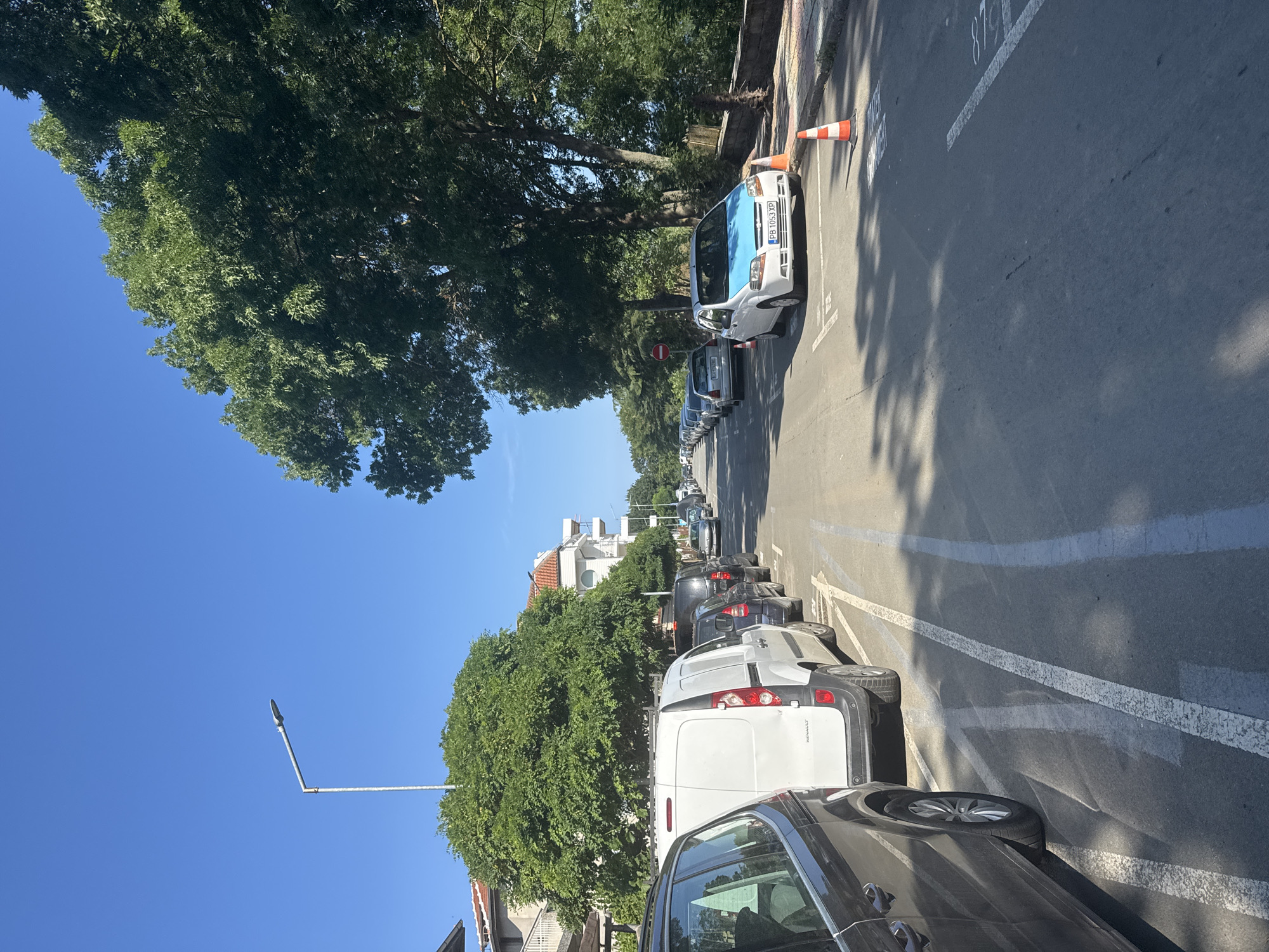 A street scene in Primorsko, Bulgaria, shows a row of parked cars along a residential street. The cars are parked on the right side of the road, parallel to the sidewalk. The street is paved and appears to be in good condition. There are trees lining the street, providing shade. The sky is clear and blue. A street lamp is visible on the left side of the image. The architecture of the buildings suggests a European setting. The overall impression is one of a typical sunny day in a small coastal town.