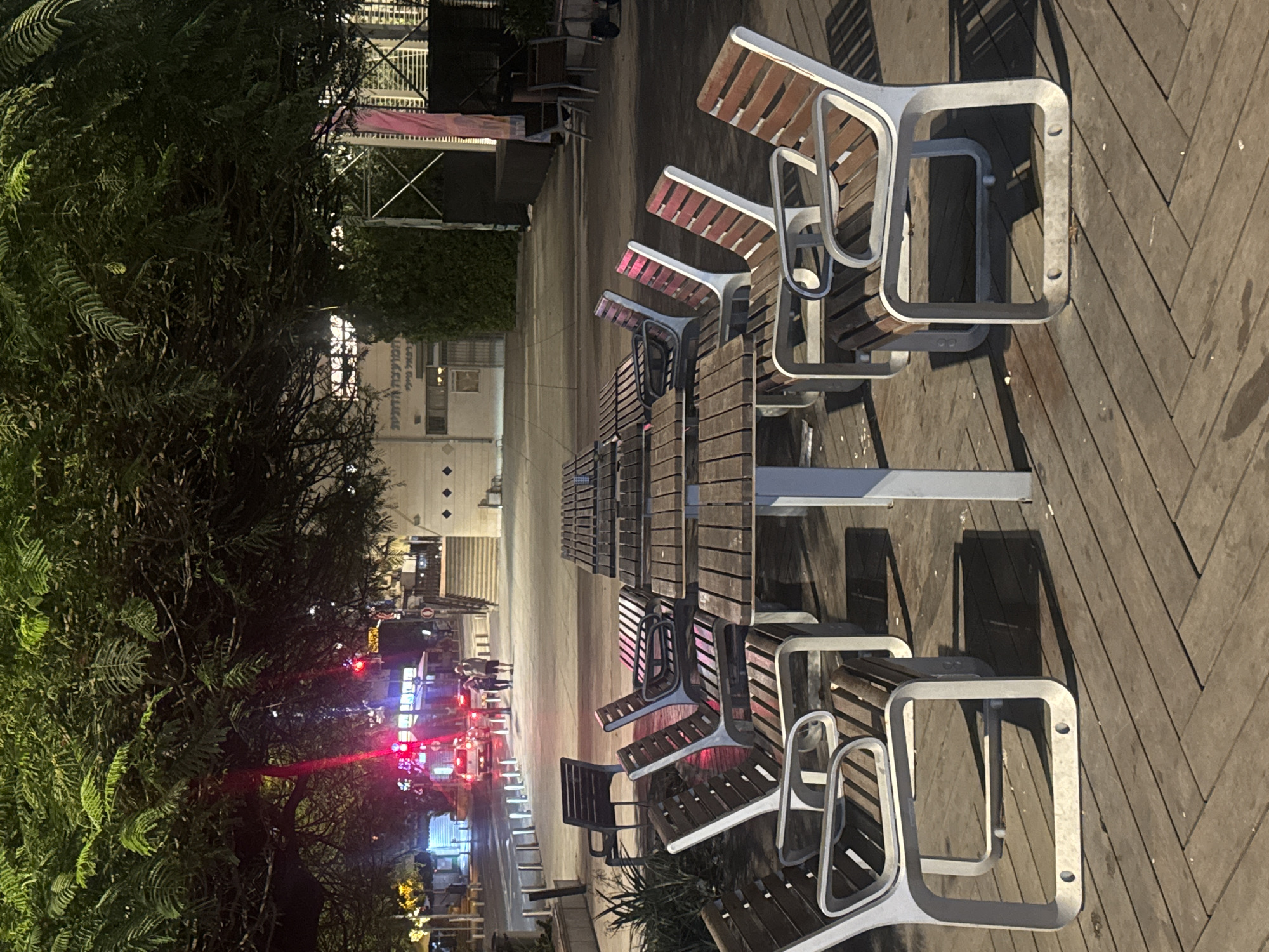 The image shows a row of modern park benches with silver frames and wooden slats situated on a herringbone-patterned wooden deck in Ramat Gan, Israel. The benches are aligned along a pathway, leading towards a street at night. Distant buildings with illuminated signs and red traffic lights indicate an urban environment. A thick row of trees borders the benches on the left side of the image. People are visible walking on the distant sidewalk.