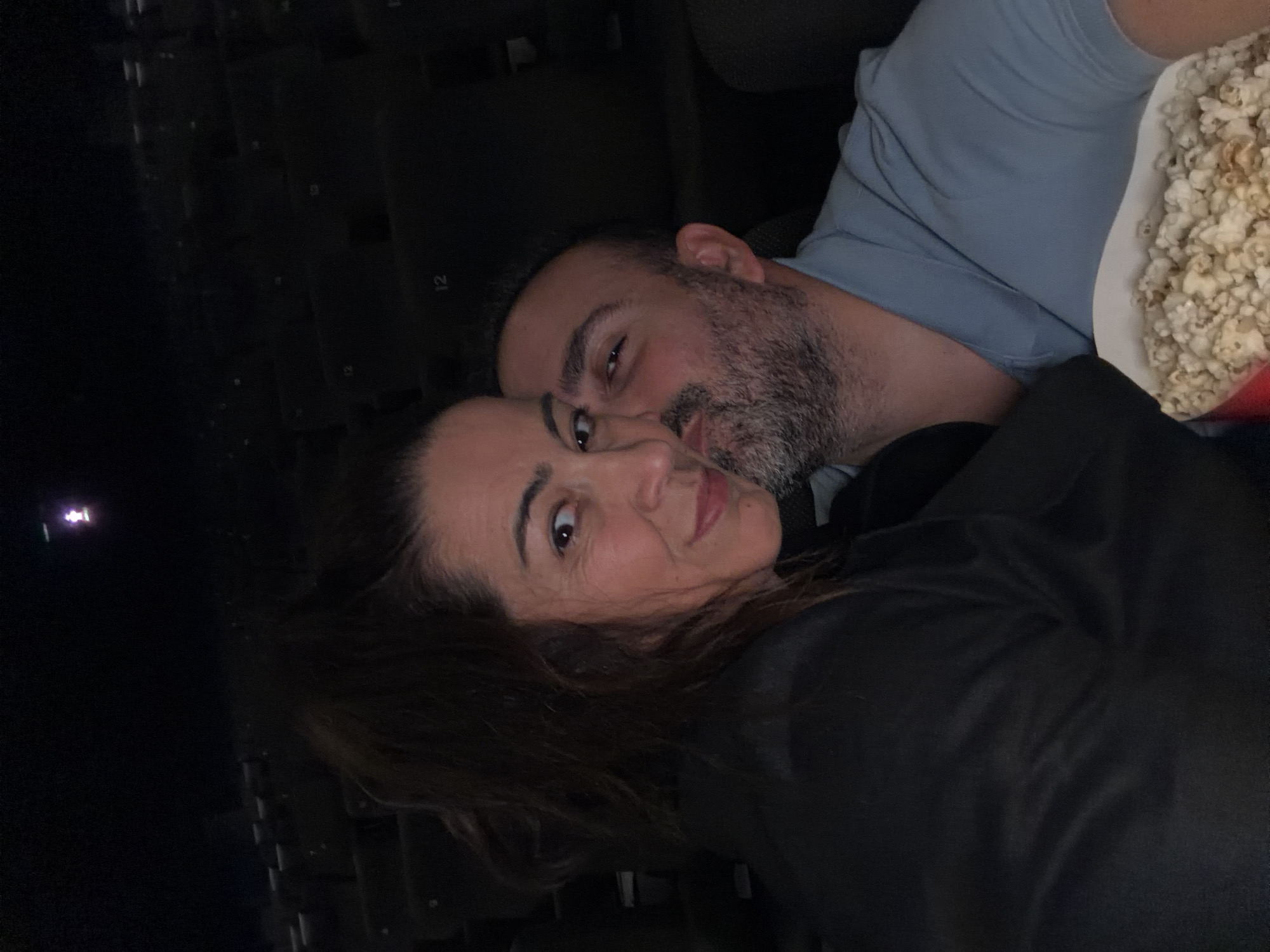 A medium shot shows a man and a woman cuddling, indoors, with a large container of popcorn in view. The man has short, dark hair and a beard. He is wearing a light blue shirt. The woman has dark hair and is wearing a black jacket. The dark background suggests the location is likely a movie theater. The presence of numbered seats in the background confirms this. The location can be further identified as a cinema in the city of Luxembourg, Luxembourg.