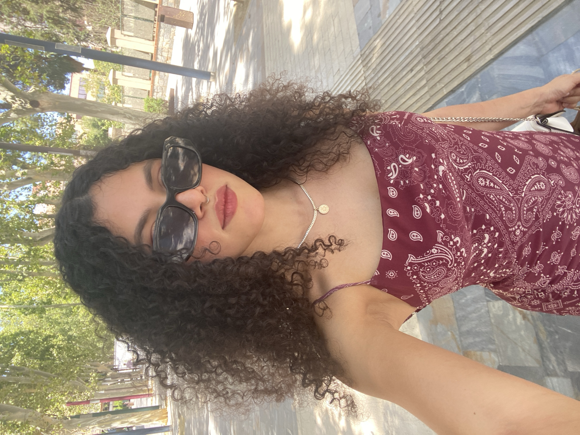 A young woman with dark, curly hair and sunglasses is taking a selfie outdoors. She is wearing a burgundy patterned dress and a necklace. She is standing on a paved walkway with trees lining the street behind her. In the background, there are buildings and a lamppost. The scene appears to be in Murcia, Spain, as indicated by the architectural style and vegetation common to the region.