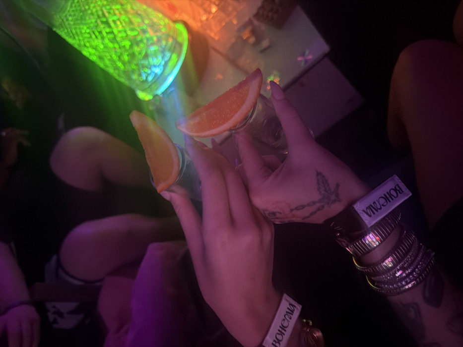 Two hands hold shot glasses garnished with orange slices in a dimly lit environment. Both wrists are adorned with numerous bangles and a wristband labeled "BOHEMIA." One hand has tattoo art. A green-lit textured object is visible above the hands. Other people are seated in the background, their legs and attire partially visible. The scene is cast in neon hues, particularly purple and pink.