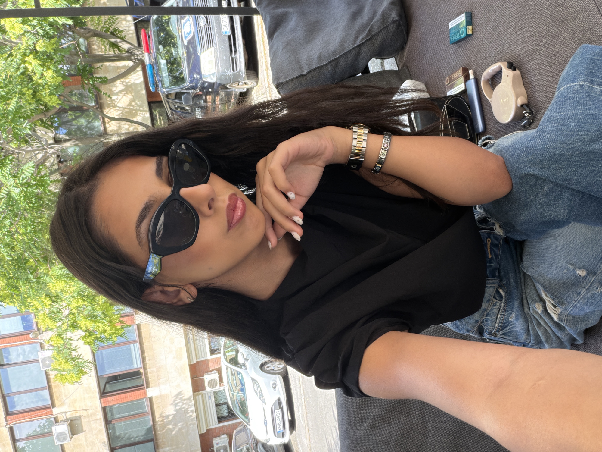In the city of Dupnitsa, Bulgaria, a young woman with long, dark hair and wearing large, black sunglasses is captured in a selfie. She is seated on a cushioned outdoor sofa, dressed in a black top and ripped blue jeans. Her right hand is raised to her chin, revealing manicured nails. She wears a gold and silver watch and a charm bracelet on her left wrist. On the surface next to her, there are two packs of cigarettes, a vaping device, and a retractable dog leash. In the background, a white car is parked on the street, and further beyond, a police van with flashing lights is visible. A building with large windows and trees with green foliage are also part of the urban backdrop.