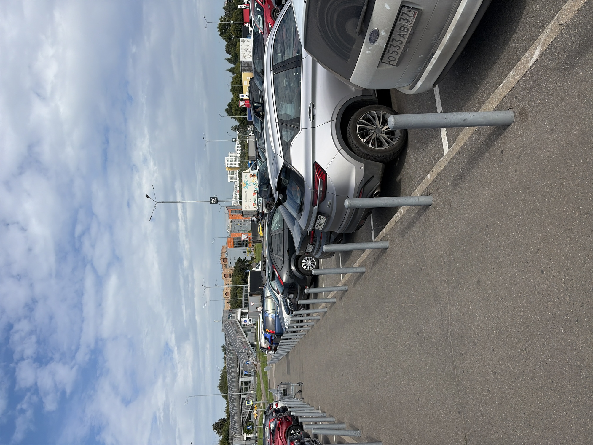The image depicts a parking lot in the city of Balashikha, Russia, likely near a commercial area, with a modern pedestrian overpass visible in the background. The parking lot is paved with asphalt and is lined with concrete bollards. Several cars are parked in the designated spaces, including a silver Geely Coolray SUV with the license plate "X50" and a silver Ford SUV with the license plate "O533AB 37". A shopping cart is positioned in the aisle between parked cars. The sky is partly cloudy. Buildings and trees are visible in the distance.
