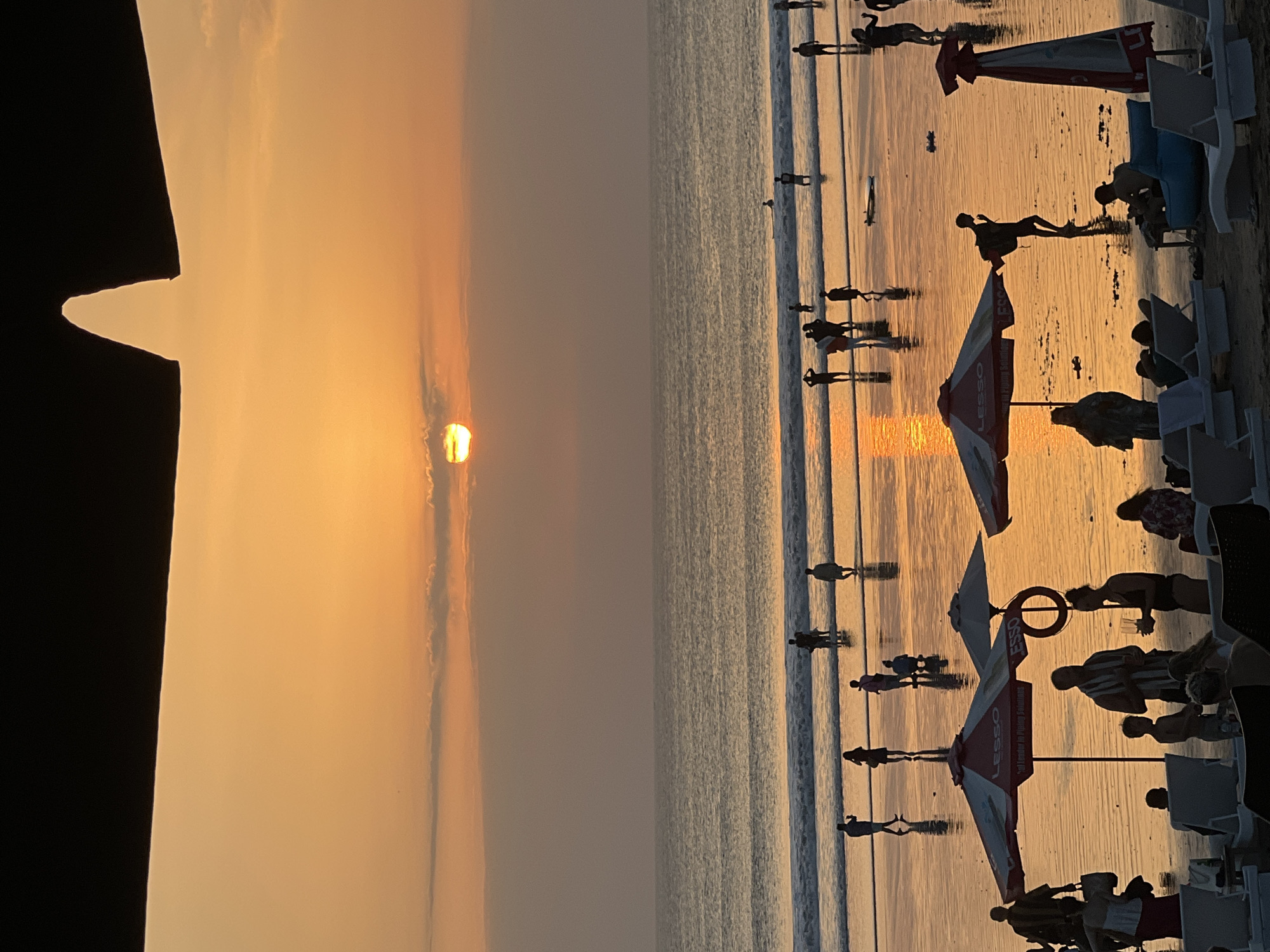 The image captures a sunset scene at a beach in Kuta, Indonesia. The sky is filled with warm orange and yellow hues, with the sun partially obscured by clouds near the horizon. The ocean stretches out, with gentle waves lapping at the shore. Silhouettes of people are scattered across the wet sand, some walking, some standing, and some wading in the shallow water. Beach umbrellas, one prominently featuring the "LESSO" logo, are set up on the sand, with lounge chairs visible in the foreground. The wet sand reflects the golden light of the sunset, creating a shimmering effect. A few people are gathered near the umbrellas, and some individuals are interacting. In the distance, a surfer can be seen on a wave, and a surfboard lies on the sand. The overall atmosphere is one of a relaxed evening at a popular beach destination.