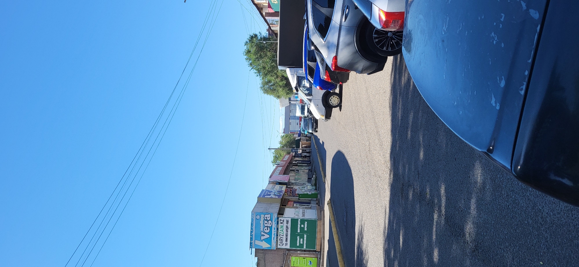 This is a ground-level, vertical, outdoor photograph taken on a clear, sunny day in Taldykorgan, Kazakhstan. The scene depicts a street lined with commercial buildings on the left, displaying various signs, including one for "Vega" and another for "QARYZZAIM.KZ". The street is paved and there are several vehicles parked along the side and in the distance, including a white van, a blue SUV, a dark SUV, and another silver car partially visible in the foreground. The environment suggests a commercial area or a marketplace. There are no people clearly visible in the frame. Overhead power lines are visible against the bright blue sky. Shadows are cast on the street from the buildings and vehicles, indicating the sun is positioned high in the sky.