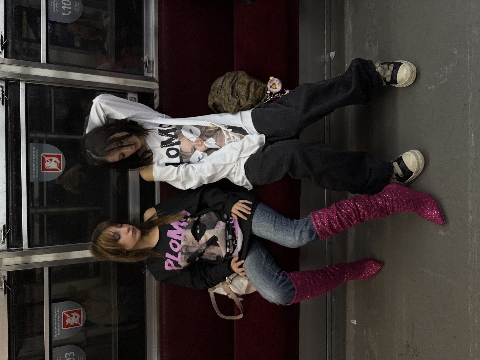 Two young women are lying down on a bench in a train car. The woman on top is wearing a white long-sleeved shirt with a picture on it and black sweatpants. The woman below her is wearing a black long-sleeved shirt with a pink design, blue jeans, and pink sparkly boots. Both women are wearing shoes. There is a backpack and a small white purse on the bench near them. The interior of the train car is visible with a maroon bench and gray floor. A window in the train car has a sticker with safety information in Spanish, indicating this is in Buenos Aires, Argentina. The OCR text also says "(10" and "ANTE U EMERGENCIA O POLICIAL LLA" which translates to "Emergency or police call".