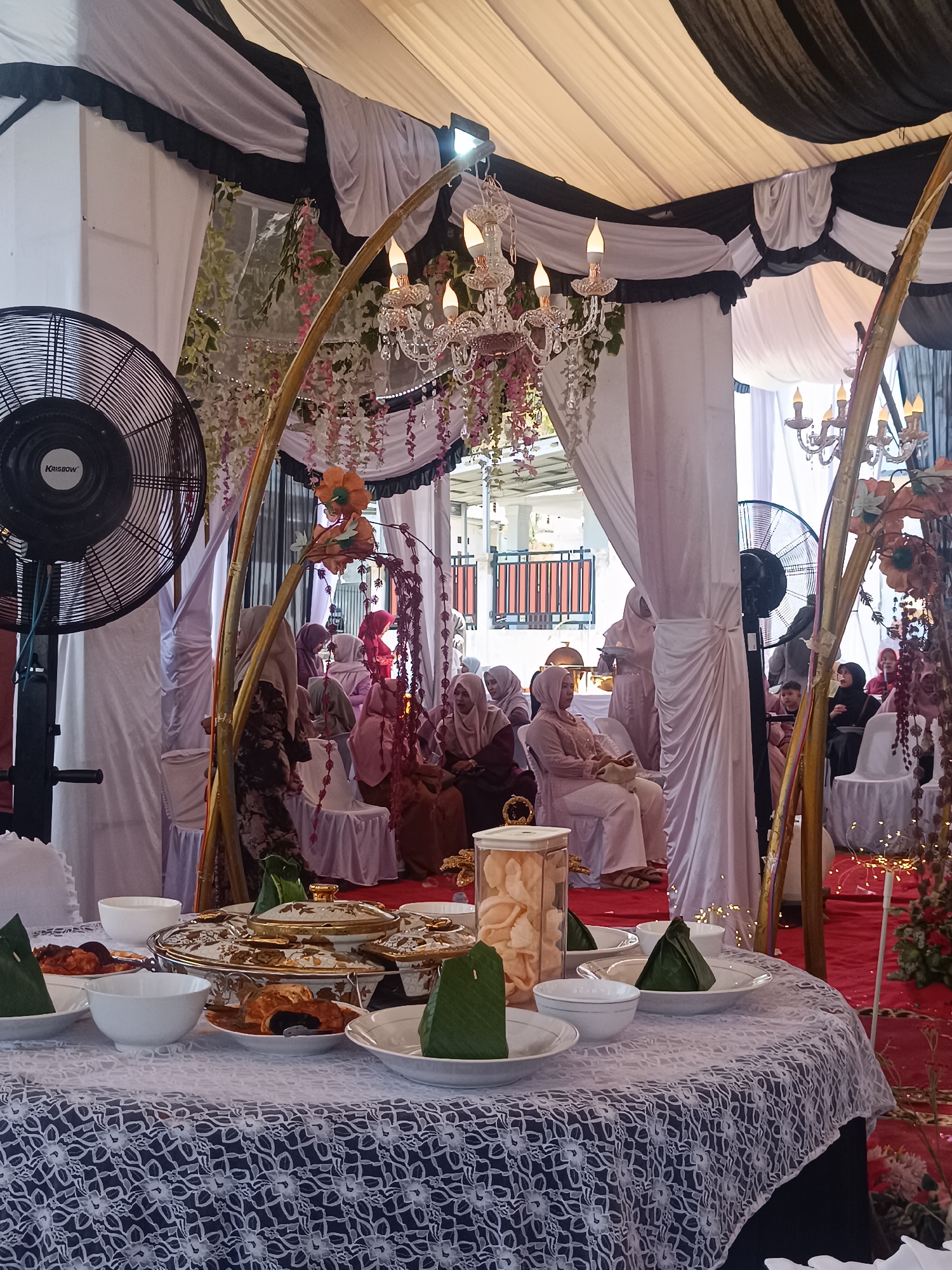 The image depicts an event occurring indoors in Lhokseumawe, Indonesia. Seated women wearing hijabs are arranged in rows, apparently waiting for the event to commence. A table is prominently displayed in the foreground, adorned with a white, lace-patterned tablecloth and various dishes, including bowls, plates with food covered in green leaves, and a container of crackers. Ornate, gold-trimmed serving dishes are also visible. The event space is decorated with draped white and black fabric, crystal chandeliers, and gold arches with floral decorations. An industrial fan is positioned in the background. A red carpet is also observed.