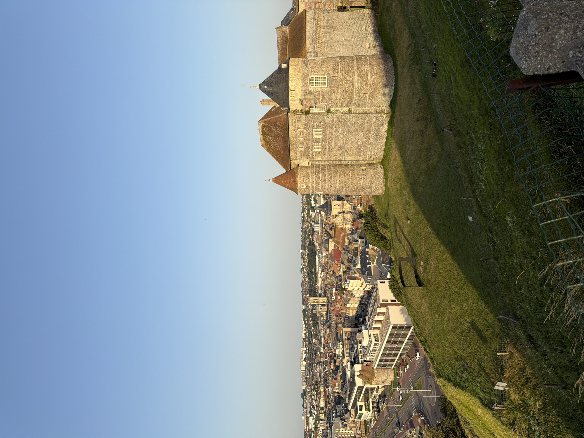 The image depicts a panoramic view of Dieppe, France, taken from a high vantage point. The scene is divided diagonally, with a vast expanse of clear blue sky occupying the left half and the town and its surroundings filling the right. Dominating the upper right portion of the frame is a large, stone castle with multiple turrets and pitched roofs, suggesting historical significance. The castle is perched on a grassy hillside, which slopes down towards the densely packed urban landscape.
The town below is a mosaic of buildings with varied architectural styles, featuring terracotta roofs and light-colored facades. A modern, multi-story building with large windows is prominently visible in the foreground of the town, contrasting with the older structures. Roads and a roundabout are visible, with a few cars suggesting some level of activity, though the overall impression is one of a quiet afternoon. The setting appears to be a coastal town, given the likely location of Dieppe.
The lighting indicates that it is either late afternoon or early evening, with long shadows cast across the grassy slope, and the sky showing a gentle gradient of color. A wire fence runs along the edge of the grassy area, partially obscuring the view of the town and the grassy slope itself. The overall mood is serene and picturesque, offering a comprehensive look at the town and its historical landmarks. There is no visible text in the image.