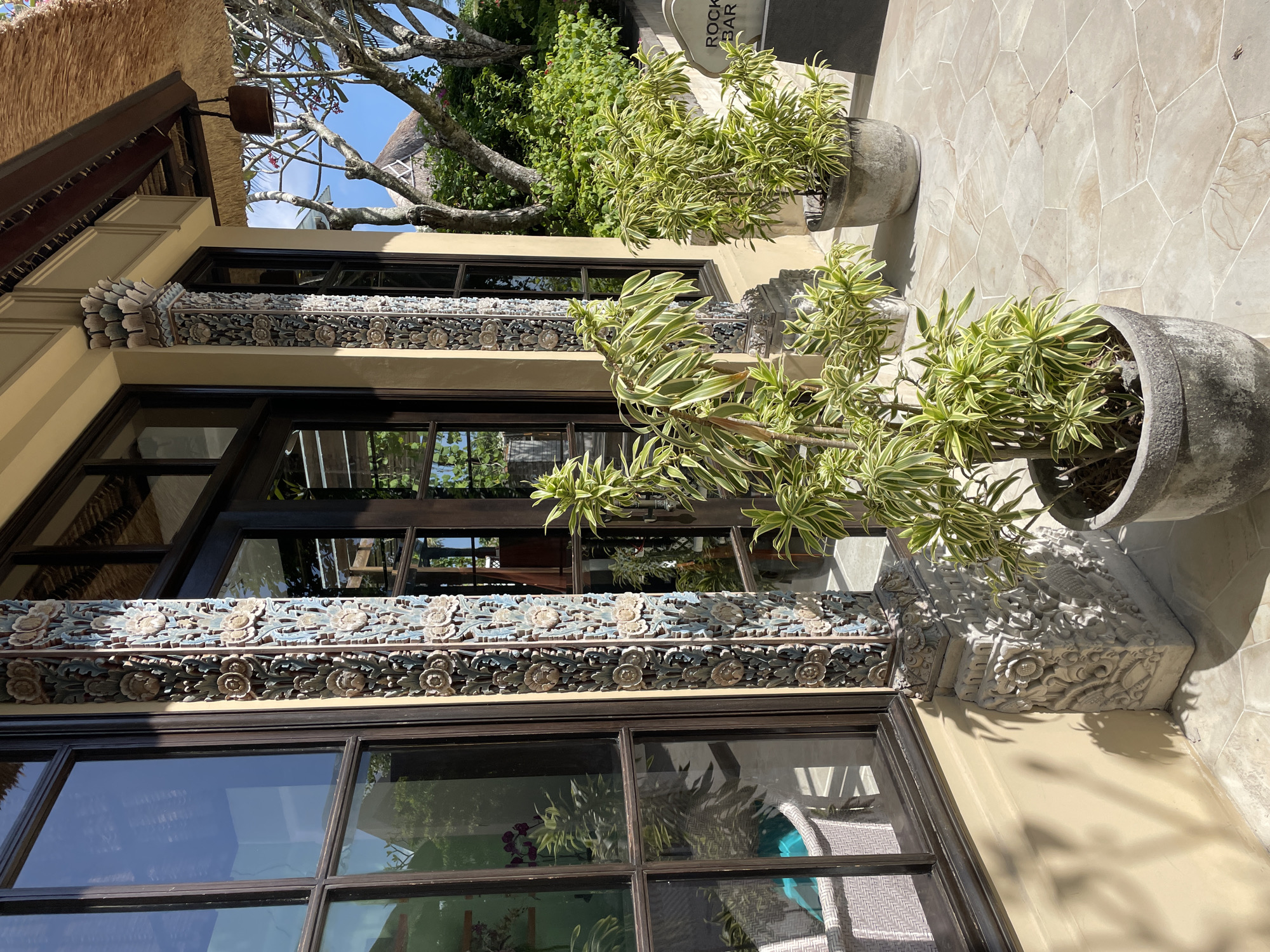 The image depicts an exterior view of a building, possibly a hotel or restaurant, located in Unknown, Unknown. The building features large windows with dark frames, decorative carved stonework above the windows, and a tan-colored exterior wall. Two potted plants with variegated foliage stand on the patterned stone patio in front of the building. A partial view of a thatched roof can be seen on the left side of the image, suggesting a tropical or resort-like setting. A sign partially visible in the background reads "ROCK BAR". The reflection in the windows shows interior details such as furniture and plants. The environment appears sunny and well-maintained.