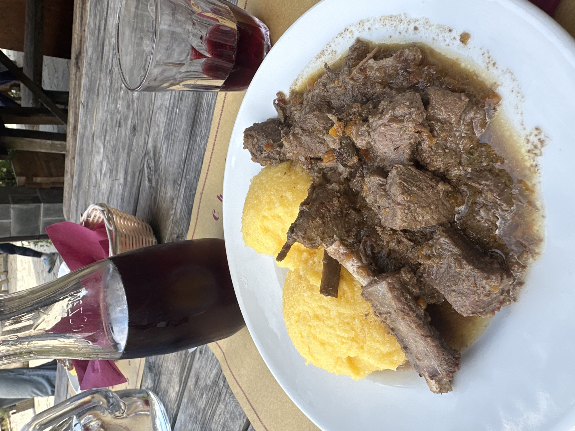 The image shows a close-up of a plate of food on a wooden table, likely outdoors at a restaurant in Corniglio, Italy. The plate contains two mounds of yellow polenta and a generous portion of braised meat in a rich sauce. The meat appears to be dark and tender, with some bones visible. To the left of the plate is a clear glass bottle filled with a dark liquid, possibly red wine or a soft drink, and a clear glass with red liquid and ice, also likely wine. In the background, a woven basket with a red napkin is visible, along with a hint of more wooden furniture and greenery, suggesting an outdoor dining setting. The overall scene depicts a rustic and hearty meal.