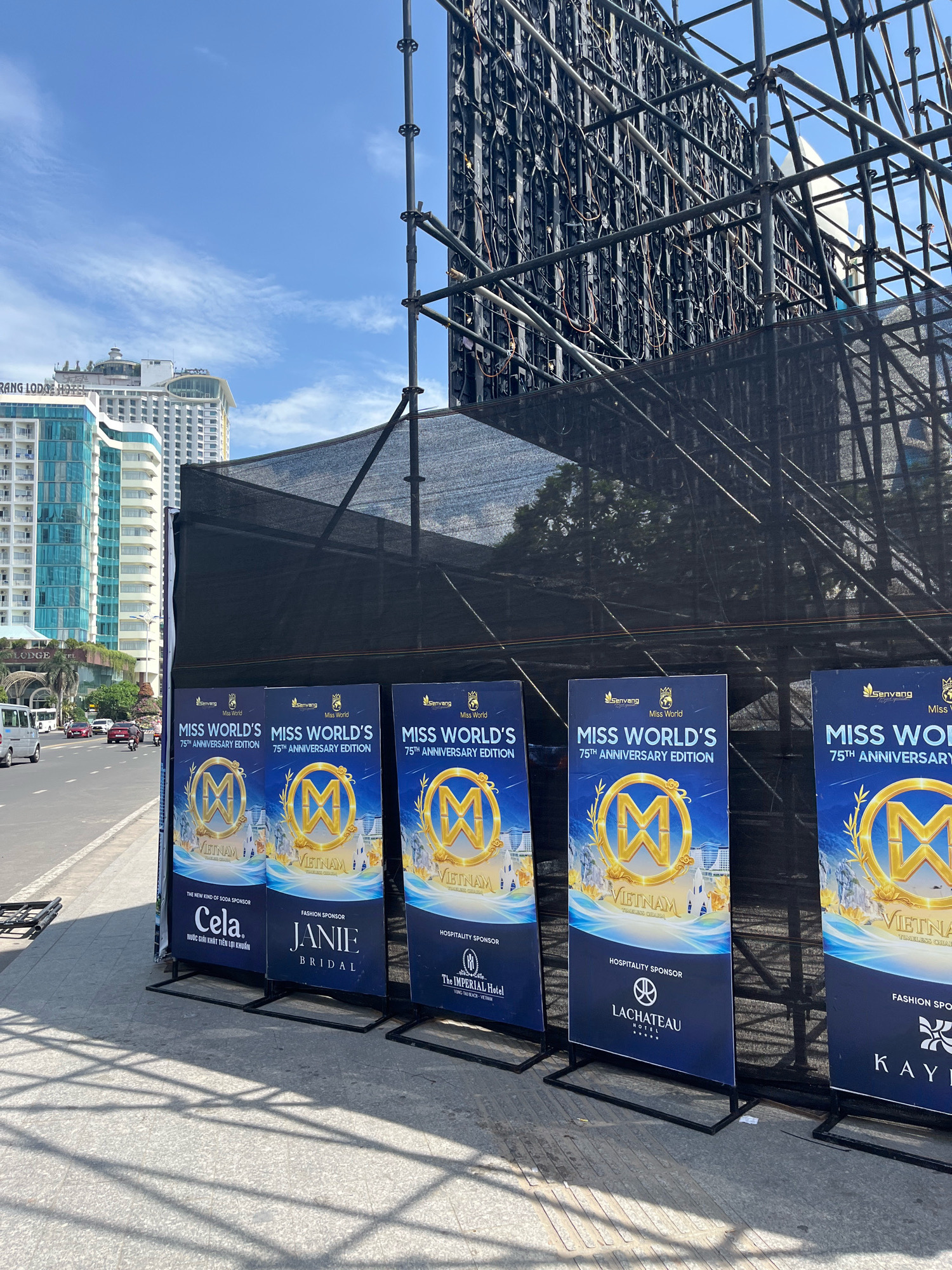 The image shows a street scene in Nha Trang, Vietnam, with a row of large blue banners displayed on the sidewalk. The banners advertise "Miss World's 75th Anniversary Edition" and feature a golden "M" logo with images of Vietnamese landmarks. Some banners also list sponsors like "Cala," "Janine Bridal," and "The Imperial Hotel." In the background, tall modern buildings and a large, dark scaffolding structure with an LED screen are visible. The street is paved, with a sidewalk and a road where cars and a motorcycle are present. The sky is blue with scattered clouds.