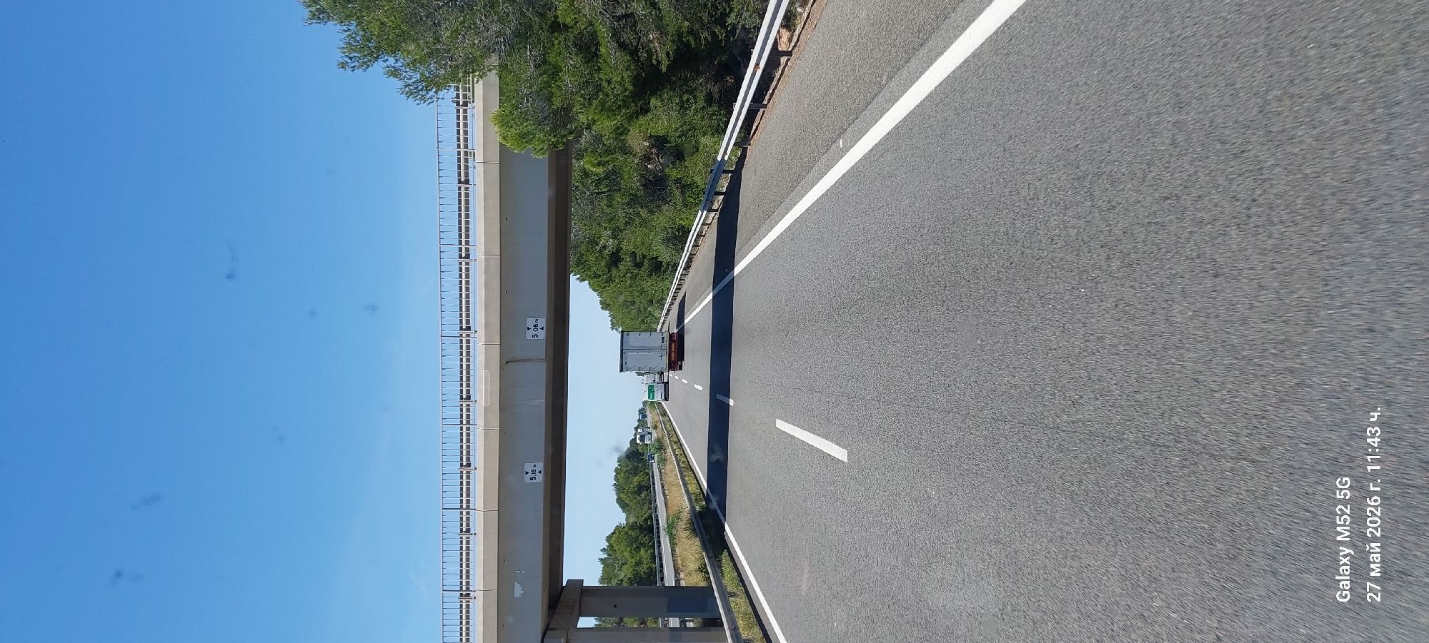 The image depicts a highway in Vandellòs i l'Hospitalet de l'Infant, Spain. The camera is oriented to the right, capturing a view of the road stretching forward. A concrete overpass with posted clearance heights of 5.06m and 5.15m spans the highway. Vehicles, including a truck, are visible in the distance on the road. The sky is clear and blue on the left side of the frame, while trees line the right side. The road surface is asphalt with white lane markings. A metal guardrail runs along the edge of the highway. Text from the camera is visible at the bottom of the image indicating "Galaxy M52 5G, 27 mai 2026 r. 11:43".