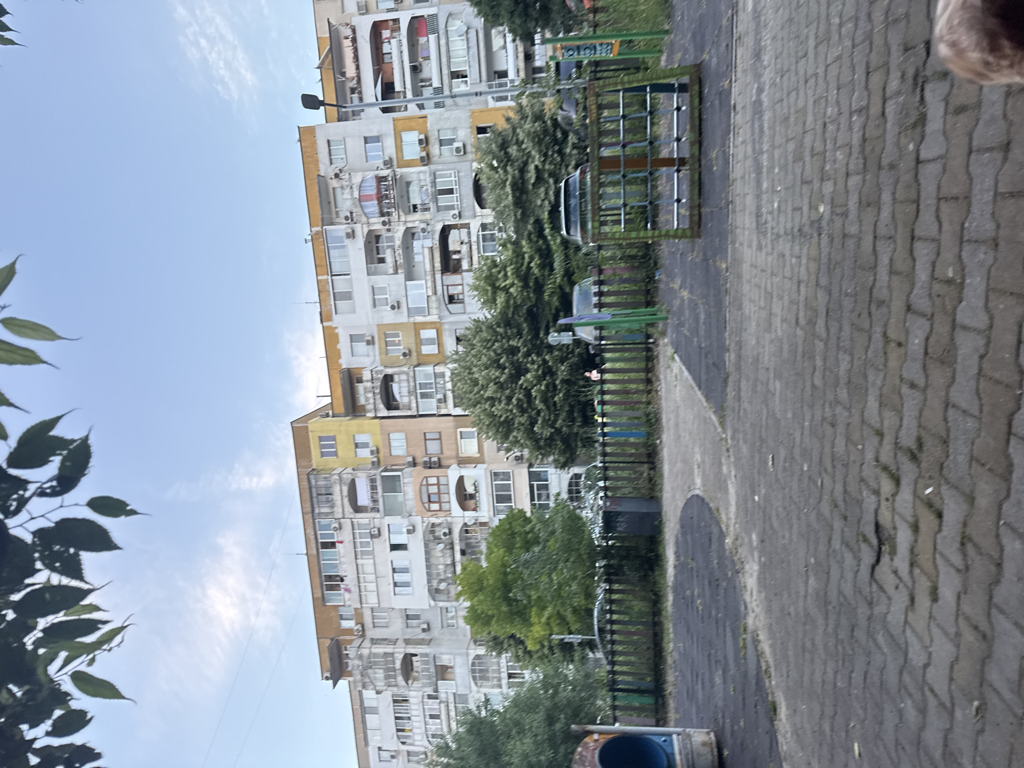The image depicts an outdoor scene in Varna, Bulgaria, focused on a multi-story apartment building. The building is weathered, with visible signs of age and varying architectural styles, including balconies and numerous air conditioning units. The facade is a mix of light gray and yellow paint. In front of the building, there's a playground area with a blue tube slide, a black trash bin, and some playground equipment with green and orange metal frames. A low, colorful fence with vertical wooden slats separates the playground from the street. Trees, some with dense green foliage, are visible behind the fence and in front of the building. A street with parked cars can be seen between the trees and the apartment building. The ground in the foreground is paved with interlocking bricks. The sky above is a clear blue with scattered wispy clouds, and tree branches with green leaves are visible in the top portion of the frame. A small portion of a brown dog's fur is visible in the bottom right corner of the image.