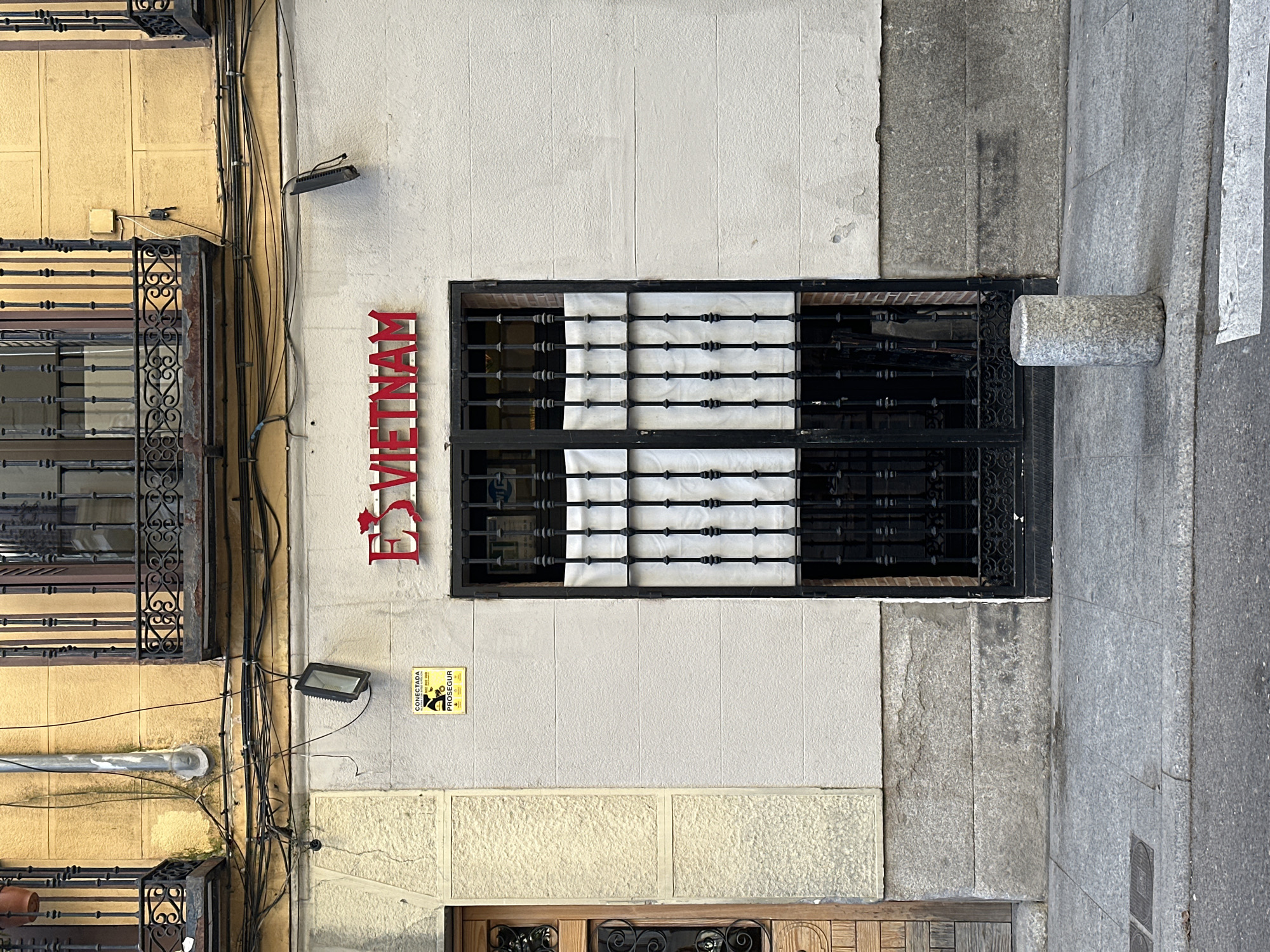The image shows a section of a building exterior. A red vertical sign spelling "ES VIETNAM" is affixed to a light-colored wall to the left of a window covered by a black metal grate with white fabric or material behind it. To the left of the sign, there is a building with ornate ironwork on its windows. There are various pipes and wires running along the wall between the two buildings. A security sign is posted on the wall below the "ES VIETNAM" sign. To the right of the window, the building material changes to a grey stone facade. Part of a street or sidewalk is visible on the right edge of the image. The presence of the "ES VIETNAM" sign suggests the location is possibly Madrid, Spain, where the Es Vietnam restaurant is located.