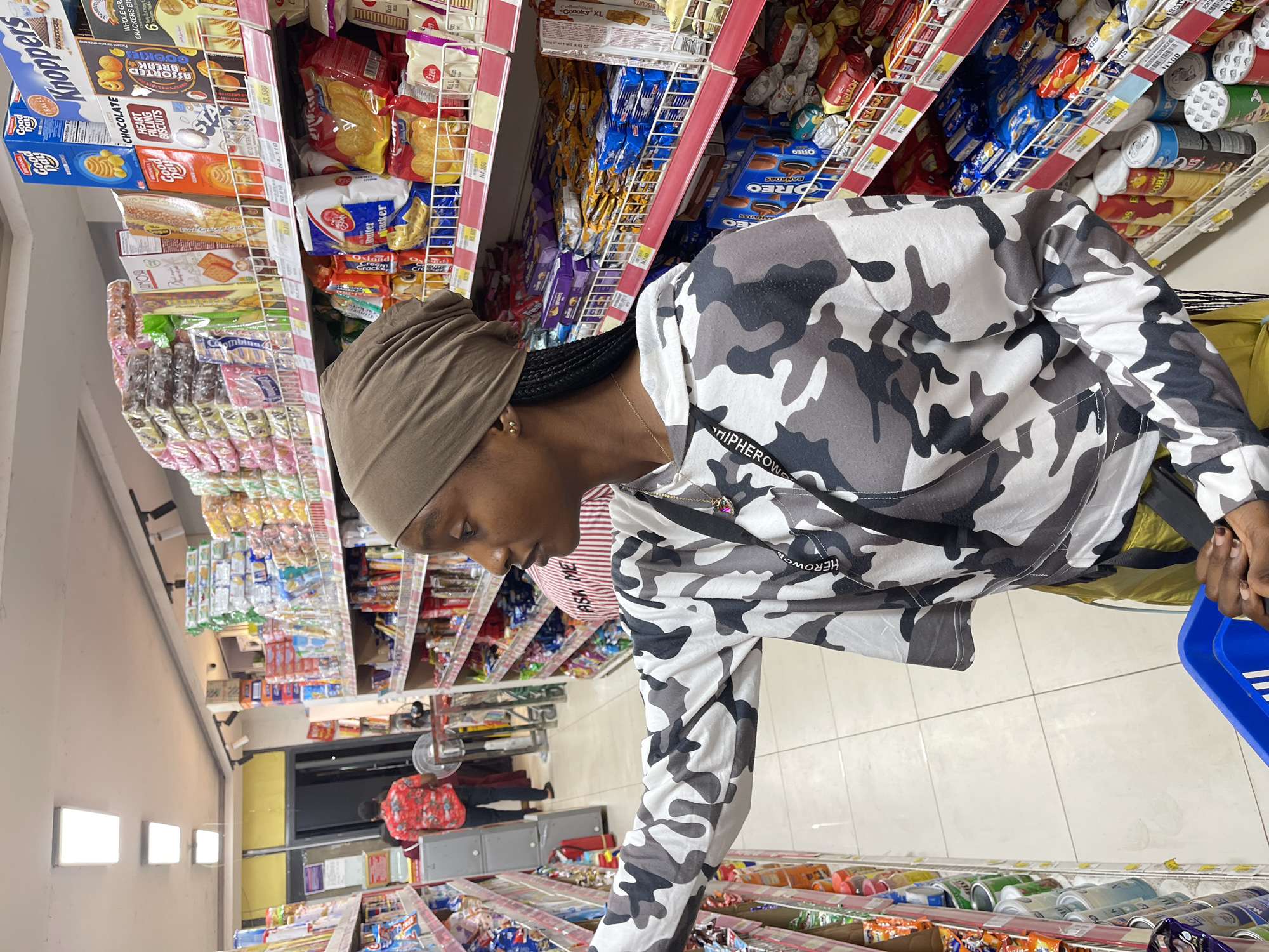 The image depicts a woman in a camouflage hoodie and a brown headscarf shopping in a well-lit supermarket aisle in Guzape, Nigeria. She is holding a blue shopping basket and appears to be reaching for items on a shelf stocked with various packaged snacks and food products, including biscuits and crackers. The shelves are organized and display a wide selection of branded goods, some with price tags in Nigerian Naira. In the background, another shopper wearing a red patterned shirt can be seen in a different aisle, and the store environment includes overhead lighting and tiled flooring. The overall impression is of a typical retail setting where customers are browsing and selecting products.
