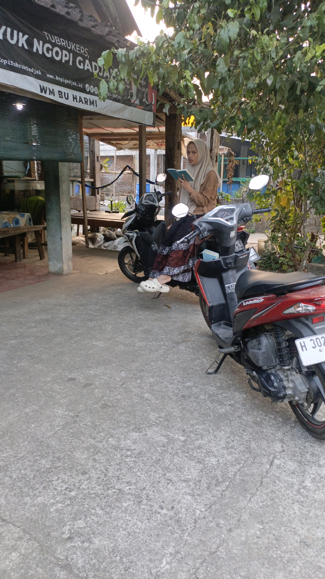 A woman wearing a hijab and a brown top is sitting on a motorcycle and reading a book. She is wearing white clogs and a patterned skirt. Behind her is another motorcycle. The scene appears to be outdoors, possibly in front of a shop or cafe with a sign that reads "YUK NGOPI GADJAH" and "WM BU HARMI." The environment is somewhat rustic, with visible wooden structures and trees. The ground is paved with concrete. The license plate of the red motorcycle in the foreground is visible and reads "H 302X." Based on the text on the sign, this location is likely in Salatiga, Indonesia, a city known for its coffee culture.