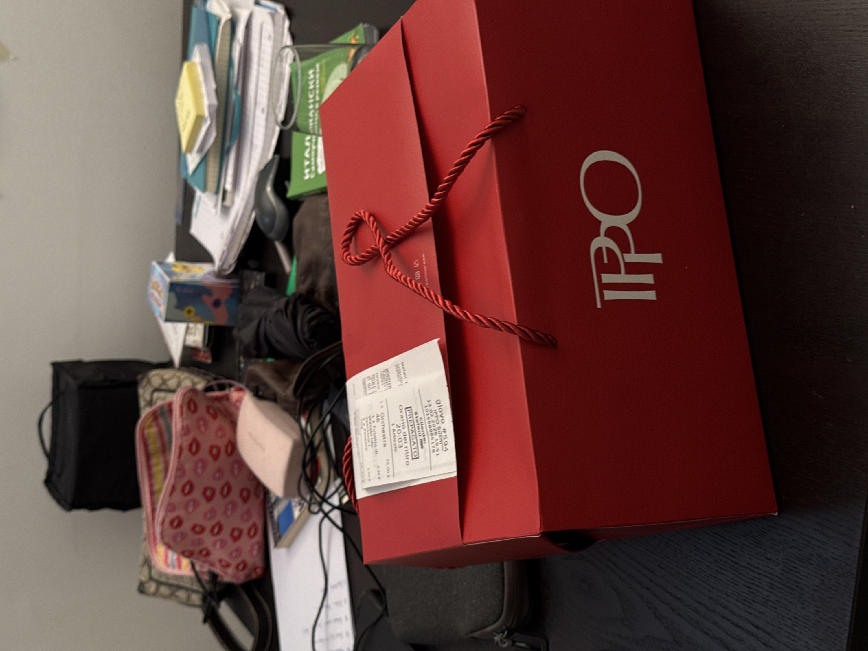 A red gift bag with "IPPO" printed in white is prominent. Attached is a receipt labeled "glovo #594 IPPO SUSHI" with a date and time of "13.02.2026 19:51," client name "Stefani," and order details including "1x Orchestra 75.00 €." The bag sits on a dark surface amid a cluttered space with a pink makeup bag, black satchel, papers, and a green box labeled with Italian text.