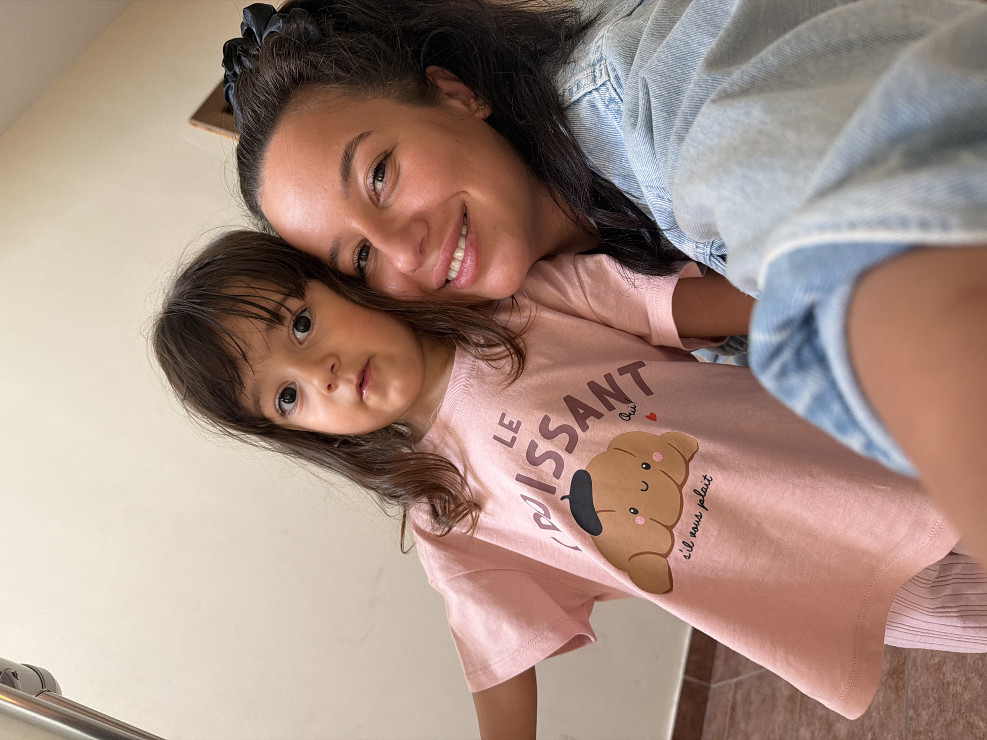 In Burgas, Bulgaria, a candid selfie captures a woman and a young girl, likely mother and daughter, posing together. The woman, with dark hair pulled back and a bright smile, wears a denim jacket. The girl, with long brown hair, sports a pink t-shirt adorned with a croissant graphic and the French phrase "Le Croissant, s'il vous plaît." They are positioned in what appears to be an indoor setting with neutral-colored walls and a hint of tiled flooring visible. The interaction suggests a moment of shared affection and playfulness.