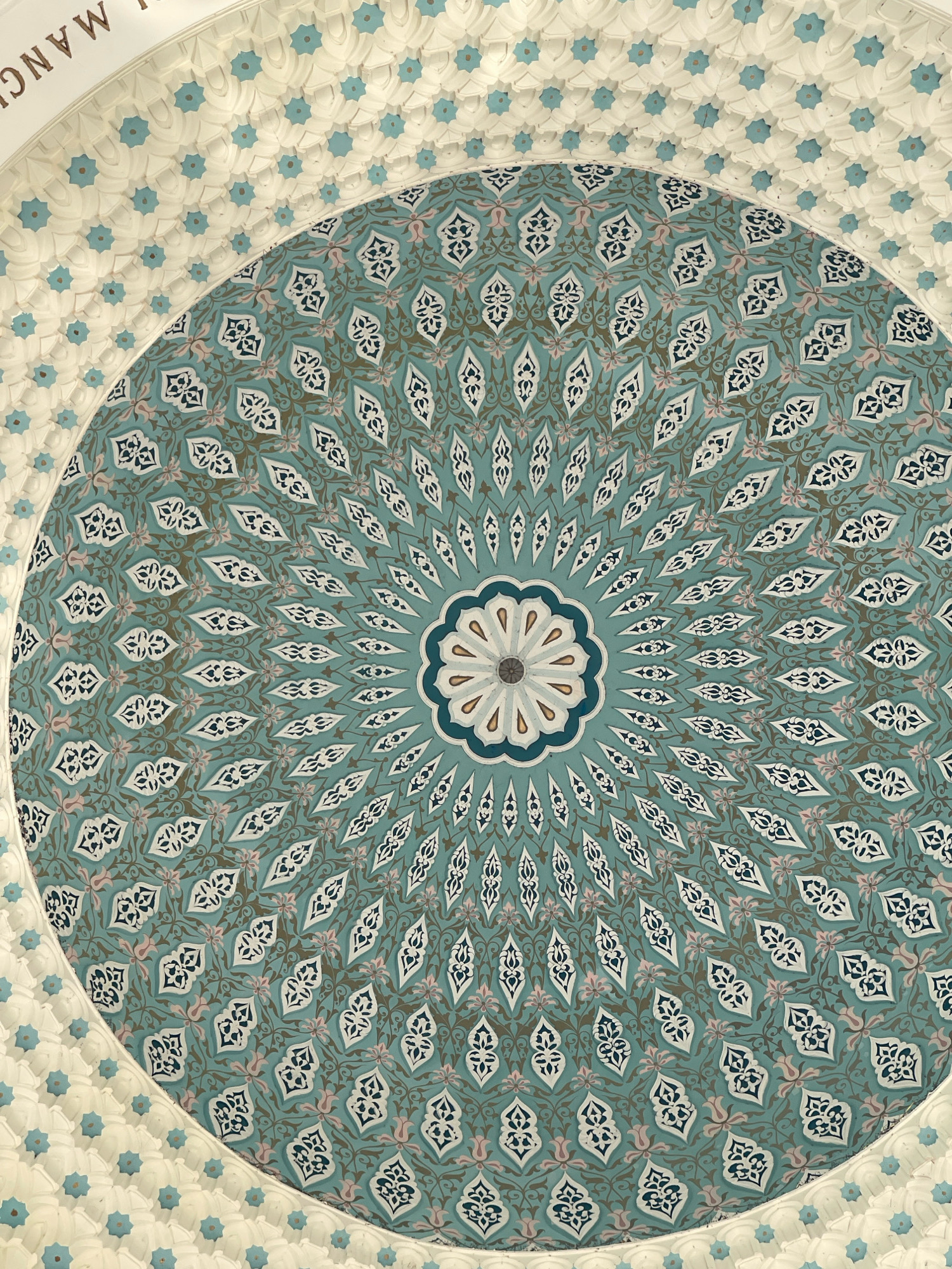 The image shows a detailed view of a decorative ceiling, likely a dome, featuring intricate Islamic geometric patterns. The central design is a stylized floral motif radiating outwards, surrounded by concentric circles of repeating paisley-like shapes and floral elements. The color palette consists primarily of teal, white, dark blue, and hints of pink and gold. The border of the dome is a textured white plaster with small blue star-shaped inlays. The overall impression is one of ornate craftsmanship and traditional artistry. The visible text "MANG" on the upper left corner suggests it may be part of a larger inscription, possibly the name of a place or person. Without further context, the specific location within Tashkent, Uzbekistan cannot be definitively identified, though such intricate decorative work is common in historical and religious buildings in the city. There are no people, actions, or interactions depicted in this static image.