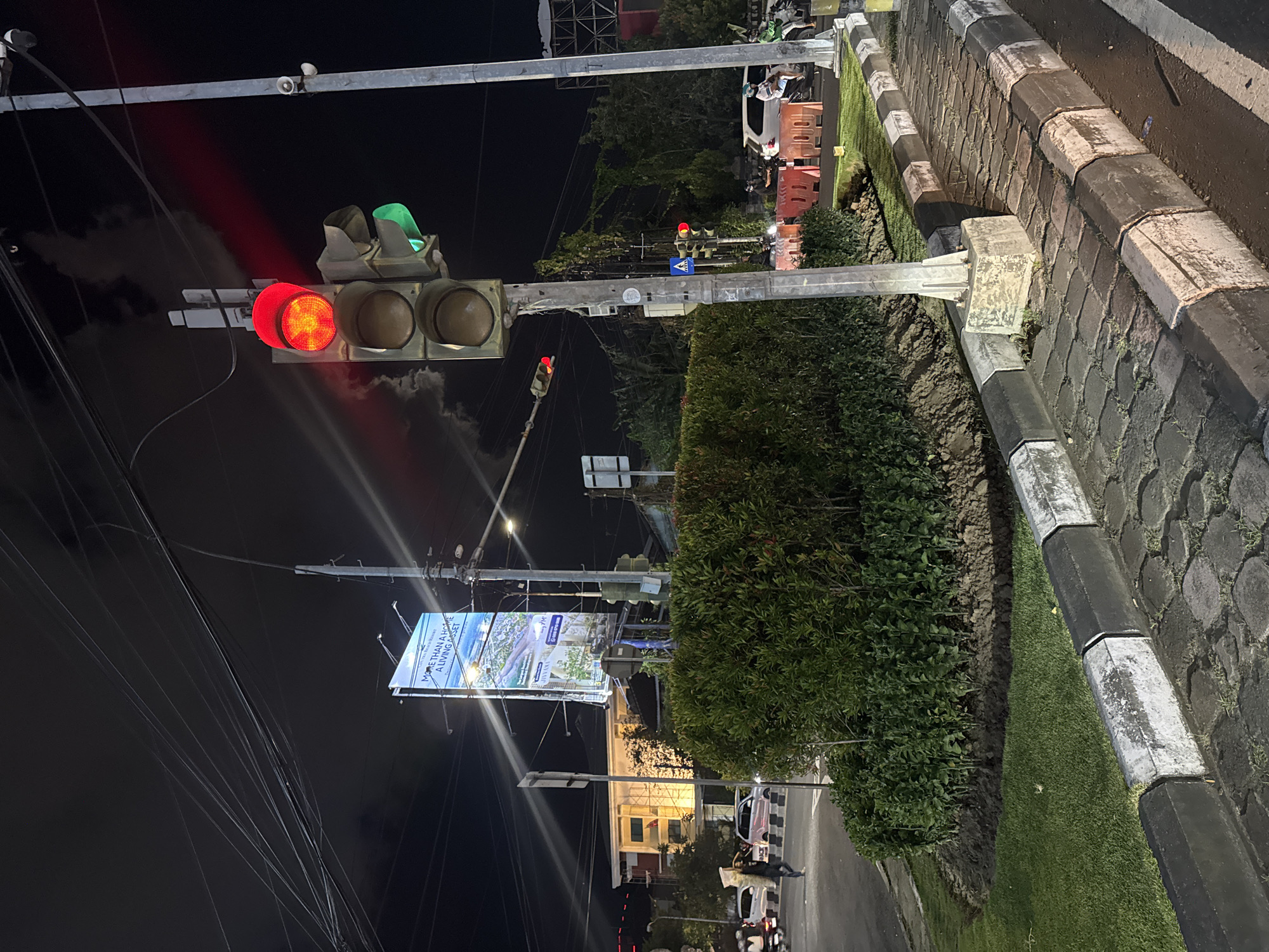 The image depicts a street scene at night in Denpasar Barat, Indonesia. A traffic light is prominently featured, with its red light illuminated, indicating a stop. The traffic light is mounted on a metal pole, which is part of a larger street infrastructure. In the background, a billboard with the text "MORE THAN A HOME, A LIVING ASSET" is visible, advertising a real estate development. The billboard features images of a modern building and a tranquil landscape.
The scene includes a paved road with a distinct curb made of stone blocks, interspersed with black and white sections. A patch of green grass runs alongside the road, bordered by dense green bushes. In the distance, trees and some buildings with lights on can be seen, suggesting an urban or suburban environment. A few vehicles, including what appears to be a white car and a motorcycle, are present on the road, indicating ongoing traffic. The sky is dark, consistent with nighttime. The overall impression is of a typical urban setting at night, with the traffic light and billboard being the most striking elements. There is no indication of specific weather conditions. The atmosphere appears functional and everyday.