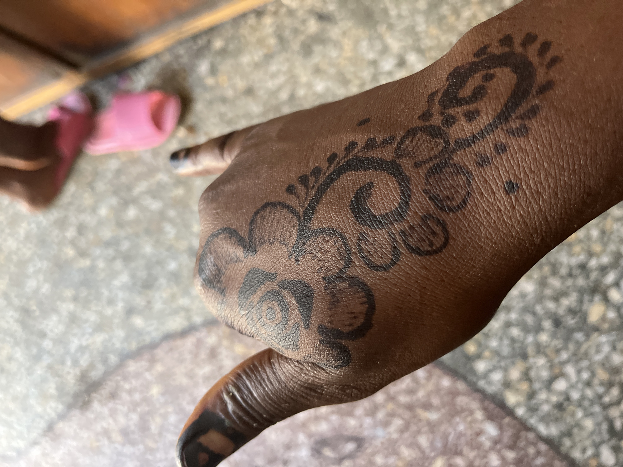 A close-up shows a dark-skinned hand with a floral henna design on the back, thumb, and fingers. The thumb and index finger are extended. The background shows a section of patterned flooring and a partially visible pink sandal near the lower left edge, close to what appears to be a wooden furniture leg. The city or specific location cannot be determined from the given image.