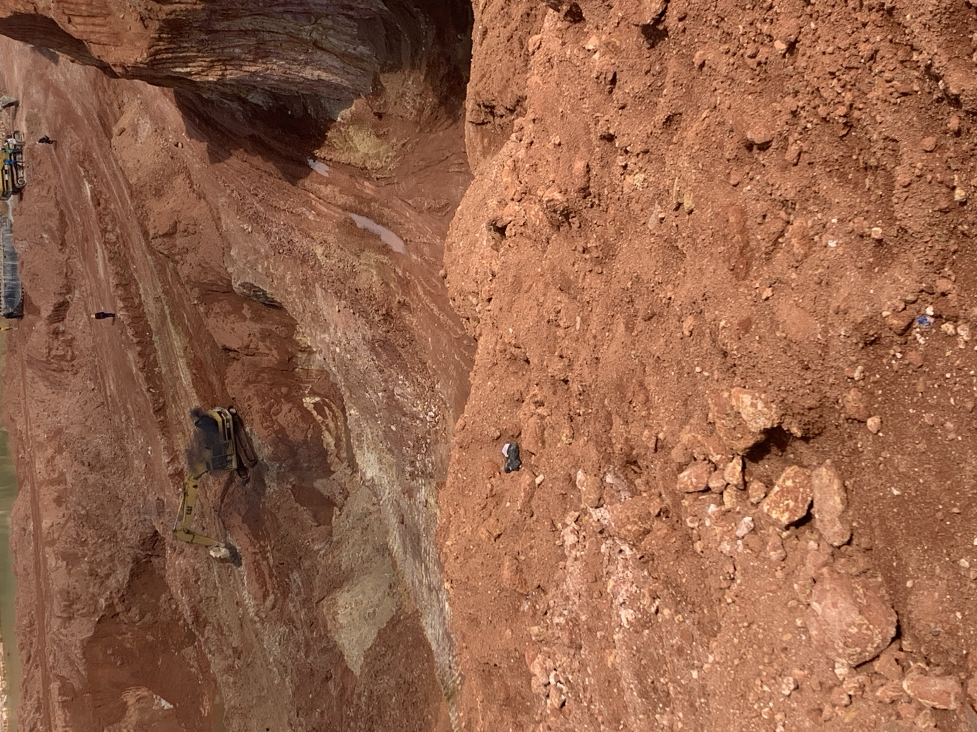 The media file shows an aerial view of a large excavation site with steep red earth cliffs. The scene is set in Kwang, Nigeria, and appears to be a quarry or mine.
The primary subjects are heavy machinery and a few individuals working at different levels of the excavation. On the left side of the image, a yellow excavator is visible, its arm extended as if actively digging or moving earth. Above it, another piece of construction equipment is partially visible. Two small figures, likely workers, are discernible on the slopes. One is near the excavator, and another is further down the cliff face. A solitary figure is also seen on the right side of the image, standing on a steep embankment. The terrain is characterized by layered red soil and rock formations, with some lighter-colored strata interspersed. Small patches of water are visible in some of the lower crevices.
The activity appears to be ongoing mining or excavation. The bright sunlight suggests it is daytime, and there are no immediate cues about the weather other than the clear visibility. The overall impression is one of industrial activity in a natural, rugged environment.
Notable details include the distinct red color of the soil, characteristic of lateritic soil common in some parts of Nigeria. The steepness of the cliffs and the presence of heavy machinery indicate a significant scale of operation. The sun's position casts shadows, suggesting it is likely midday or early afternoon.
No visible text is present in the image.
