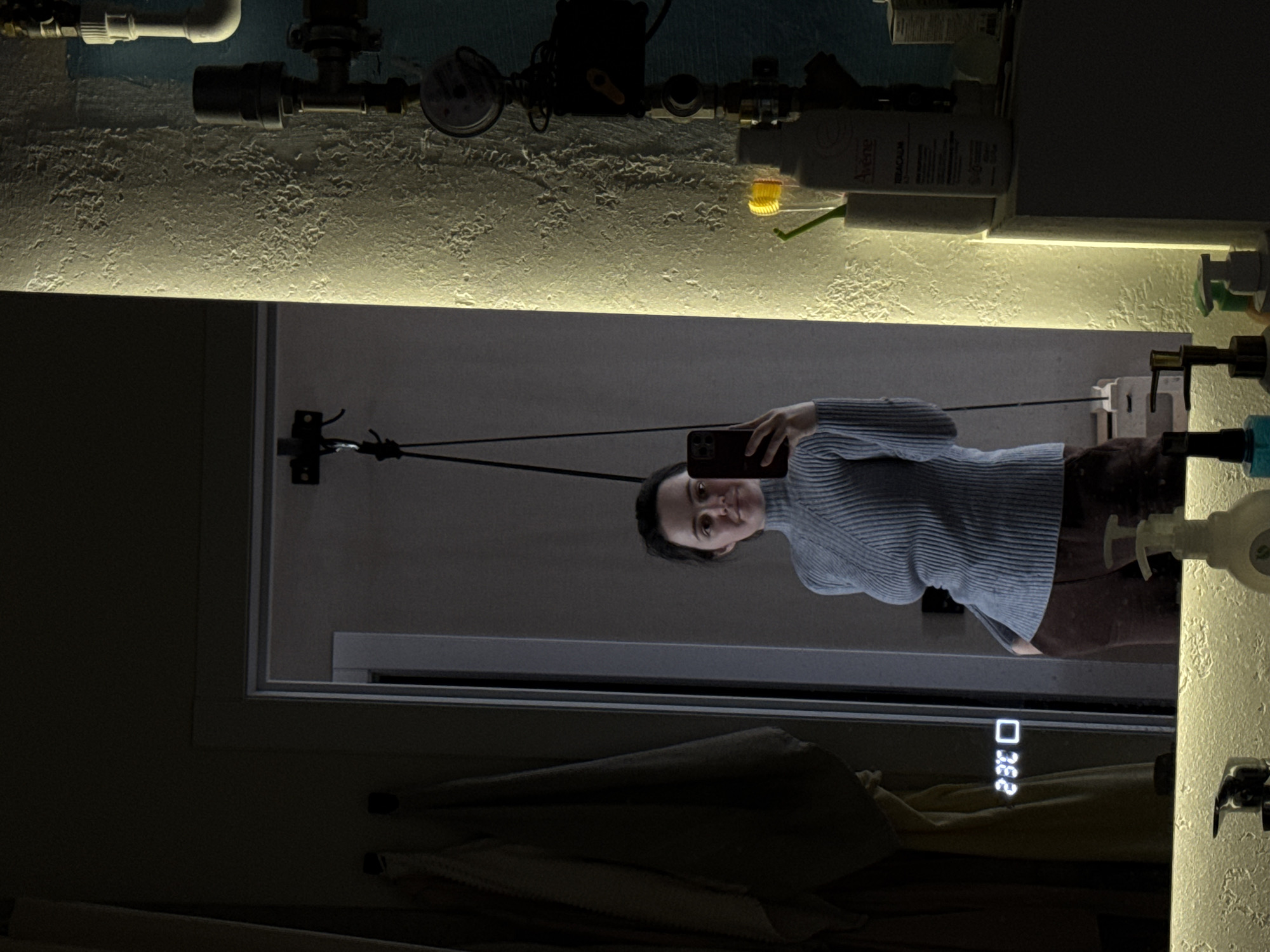 The image depicts a person reflected in a mirror within a bathroom. The person, a young woman with dark hair, is holding a smartphone and taking a selfie. She is wearing a light gray turtleneck sweater and a brown skirt. The bathroom has a lighted mirror with a digital display showing "36.2". Above the mirror are pipes and tubes attached to the textured wall. There are also toiletries and a toothbrush. Beneath the mirror, a towel or garment is hanging, obscuring the lower part of the reflection. There is no information about the location being Izhevsk, Russia, in the image.