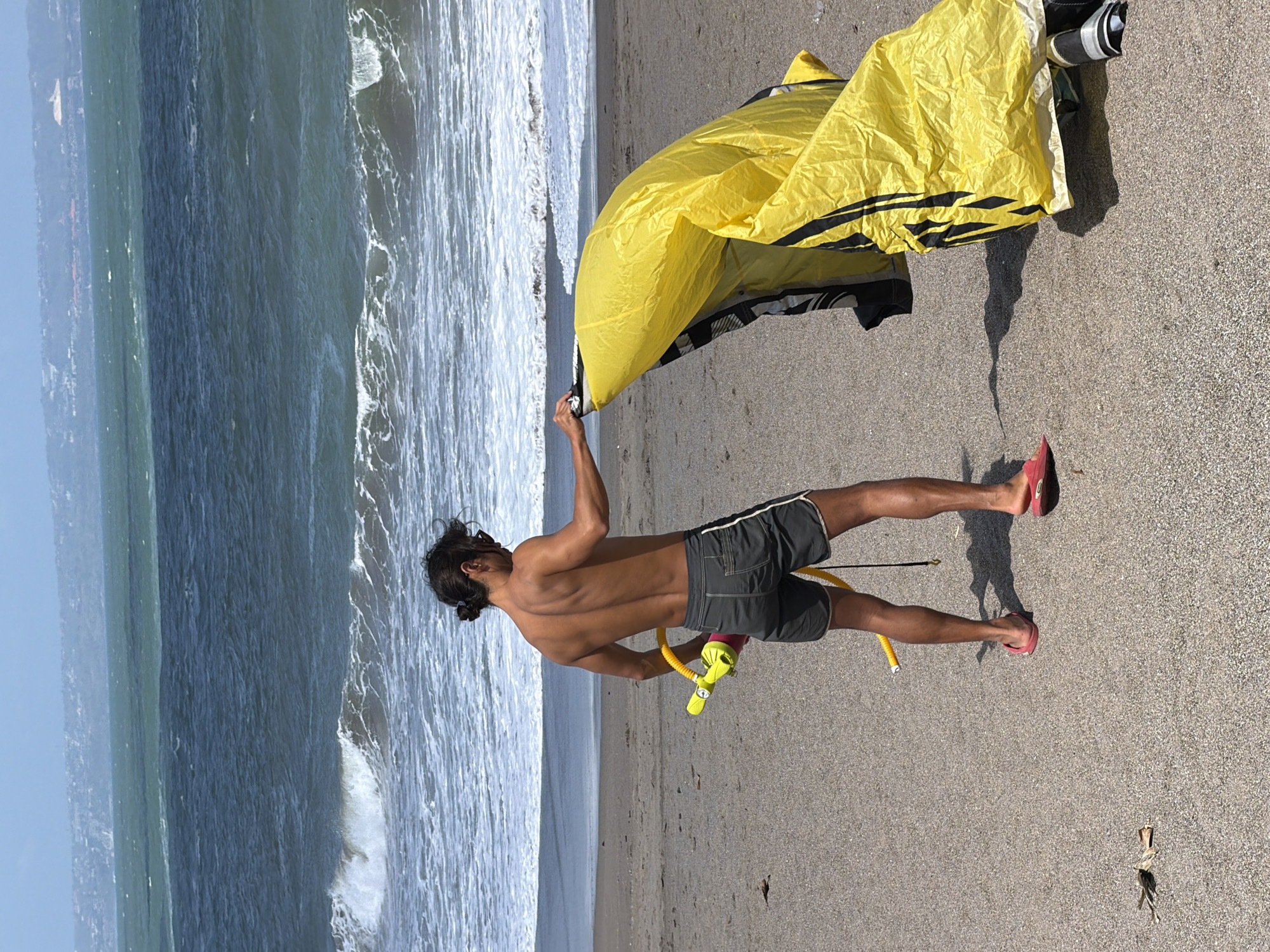 A bare-chested person with dark hair tied back in a bun stands on a sandy beach in North Kuta, Indonesia, facing away from the camera and towards the ocean. The person is wearing dark shorts and pink flip-flops. They are holding a yellow kite and a yellow pump with a pink handle. The ocean has moderate waves and is a deep blue color with a lighter green band further out. The sky is bright blue with some hazy clouds. The beach sand is a light brown color.