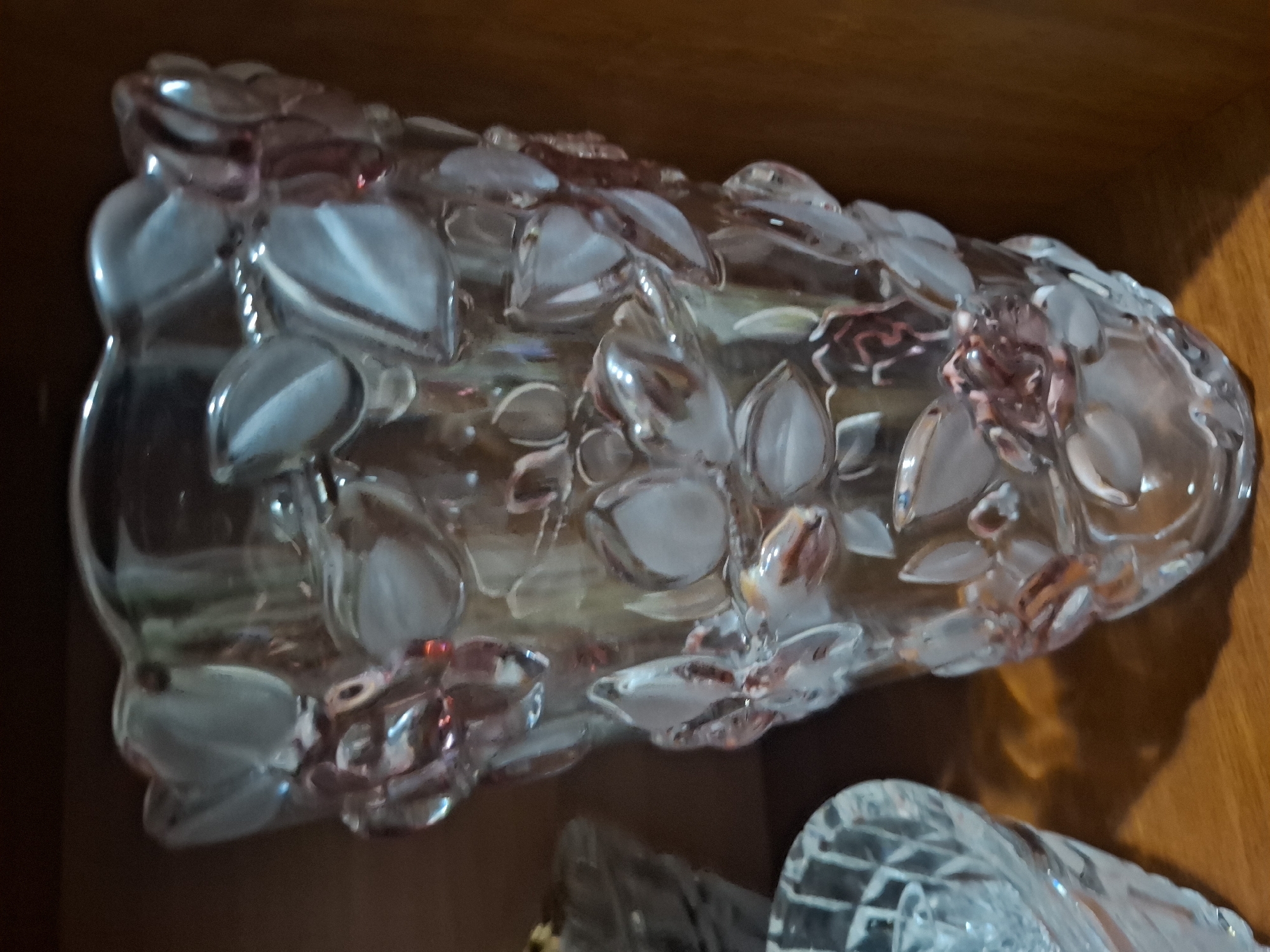 A close-up, eye-level shot shows a clear glass vase with raised floral and leaf patterns. The vase has a flared rim and is filled with an indistinct, translucent substance. It is placed on a wooden surface, likely a shelf, with a darker wooden background behind it. To the left of the vase, a portion of another clear glass object with a diamond-cut pattern is visible. The lighting is soft, creating subtle shadows and reflections on the glass surfaces. The image is not taken in Almaty, Kazakhstan.