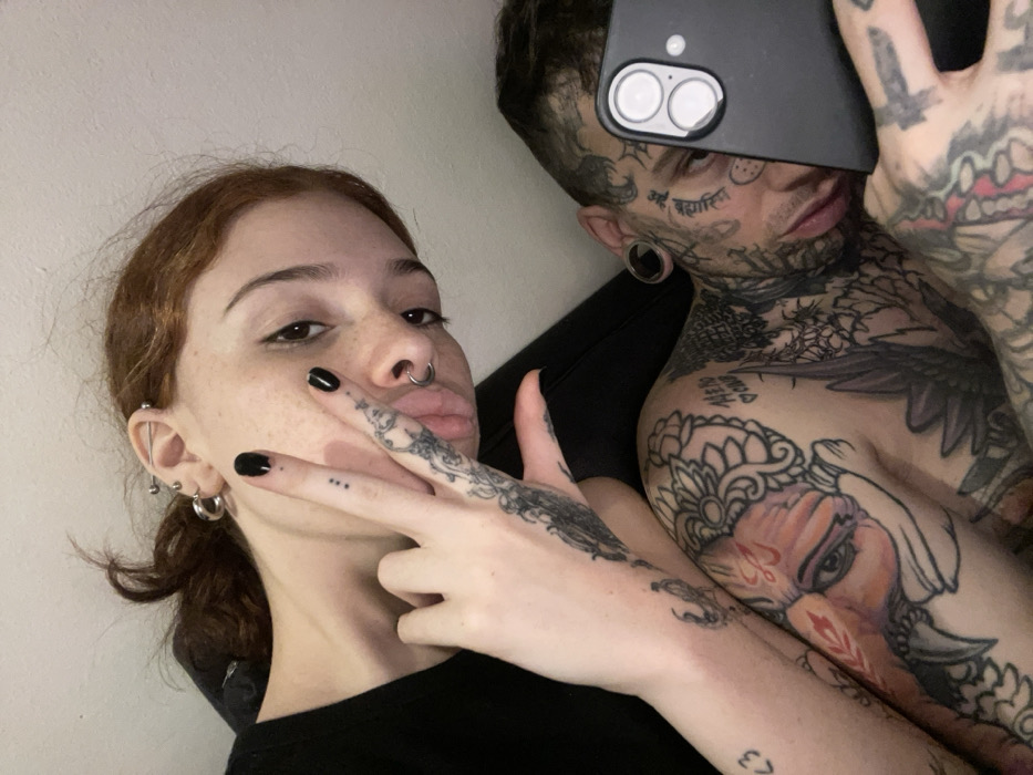 This image captures two individuals, a female and a male, in an indoor setting within the city of Buenos Aires, Argentina. The background consists of a plain, light-colored wall above a dark, possibly upholstered, surface.
The female, positioned on the upper left, has reddish-brown hair, a light complexion with visible freckles, and dark eyes with eyeliner. She exhibits multiple ear piercings, including an industrial bar and several hoops, and a silver septum ring. Her left hand is raised, forming a V-sign with the index and middle fingers. Both fingers and the back of her hand are adorned with intricate black ink tattoos, and her nails are painted black. A small three-dot tattoo is visible on her middle finger. She is wearing a black top.
The male, positioned on the lower right, features extensive black and color tattoos covering his visible right shoulder, chest, neck, and face. Facial tattoos include script/symbols on the cheek and forehead, and a spider web design near his right temple. Both earlobes display large black ear gauges. A prominent colorful Ganesha-like tattoo is visible on his left shoulder/bicep, and a heart with "ME NO CLUB" text is tattooed on his chest. He holds a black smartphone vertically with his left hand, partially obscuring his face; the phone's dual-lens camera module and flash are clearly visible. His left shoulder is positioned adjacent to the female's arm, which rests upon it. Both individuals are looking towards the camera.