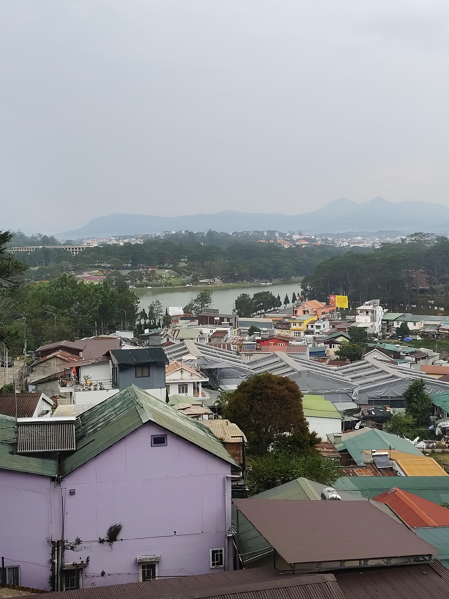 A high-angle view captures a section of Da Lat, Vietnam, under an overcast sky. A lake surrounded by trees and buildings occupies the midground, leading to distant mountains obscured by haze. The foreground displays a dense collection of rooftops in varying colors and materials, with a prominent lavender building in the left foreground. The scene is dominated by the built environment interspersed with green vegetation. No people are visible in the frame.