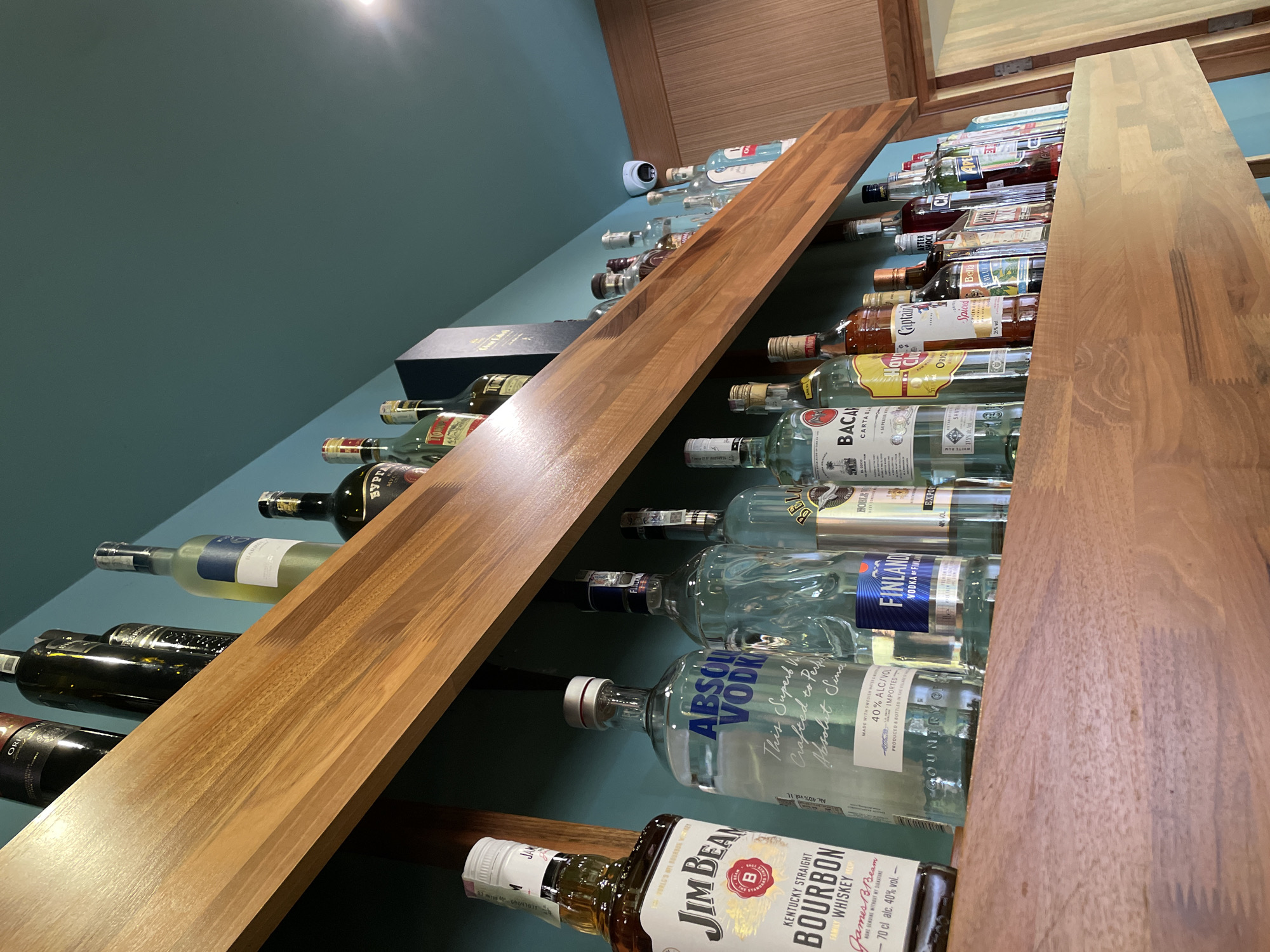 This image presents an indoor scene featuring a well-stocked display of various alcoholic beverage bottles. The setting appears to be a bar, restaurant, or a dedicated home bar area. The background is a muted teal-green wall, serving as a contrasting backdrop for the wooden shelving system.
Numerous bottles of spirits are arranged horizontally, some stacked two or three deep, between two prominent wooden planks that act as shelves or dividers. These shelves are made of light-to-medium brown wood with a visible grain. The bottles showcase a diverse selection of brands, including Jim Beam Kentucky Straight Bourbon Whiskey, Absolut Vodka, Finland Vodka, Bacardi Carta Blanca Rum, Captain Morgan Spiced Rum, After Shock, and Bell's. A black box, potentially for a "Black Label" product, is partially visible on one shelf.
Visible text includes:
- On Jim Beam: "KENTUCKY STRAIGHT BOURBON WHISKEY", "James B. Beam", "NOKE GENUINE WITHOUT MY SIGNATURE", "-70 cl alc. 40% vol. -"
- On Absolut Vodka: "This Superb W Crafted to Per Absolut Sina", "40% ALC/VOL", "IMPORTED"
- On Finland Vodka: "VODKA OF FINLI"
- On Bacardi: "BACA CARTA BL"
- On Captain Morgan: "Spiced"
Other discernible partial texts include "ORIG", "AFTER SHOCK", "Bell's", "Blu", and "Burga".
A small white security camera or sensor is mounted on the teal-green wall in the upper right section of the frame. The image is taken from a low-angle perspective, looking up at the bottles. The lighting is artificial, with a bright source visible in the upper left corner. There are no people present, and no specific activity is taking place, emphasizing the display itself. The location context provided is Nesebar, Bulgaria.