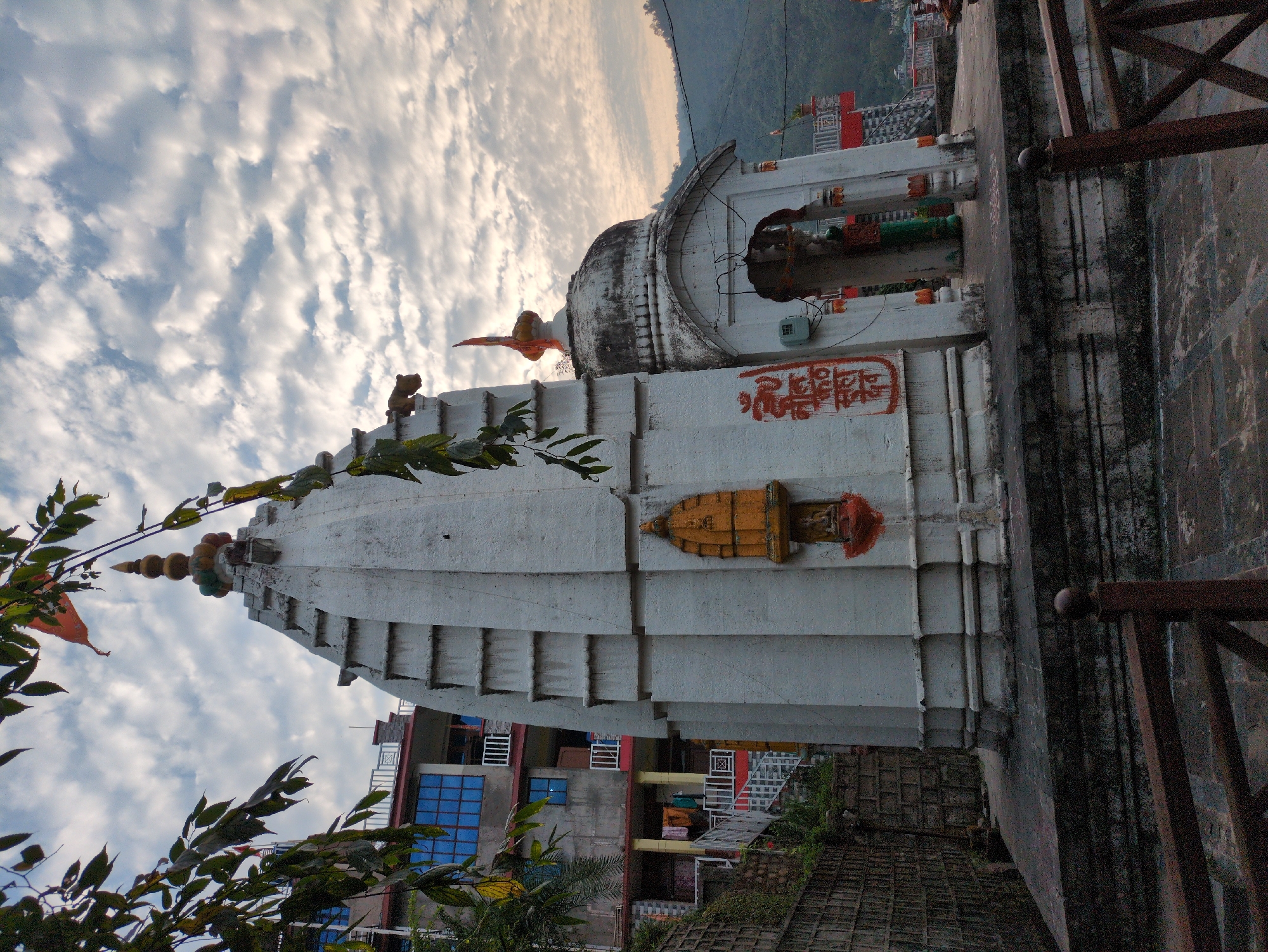 This is a low-angle, full shot of the Bhootnath Temple in Mandi, India, taken on a cloudy day. The temple, constructed from white stone, features a tiered shikara topped with decorative finials. To the right of the main structure is an arched entranceway with a dome. Orange script is painted on the wall to the left of the entrance. In front of the temple is a paved courtyard with a low stone wall and a wooden railing. A modern building with blue windows is visible behind the temple. Lush green foliage frames the top left corner of the image. The surrounding environment appears to be hilly, with visible trees and power lines in the background. The scene is static and no people are visible.